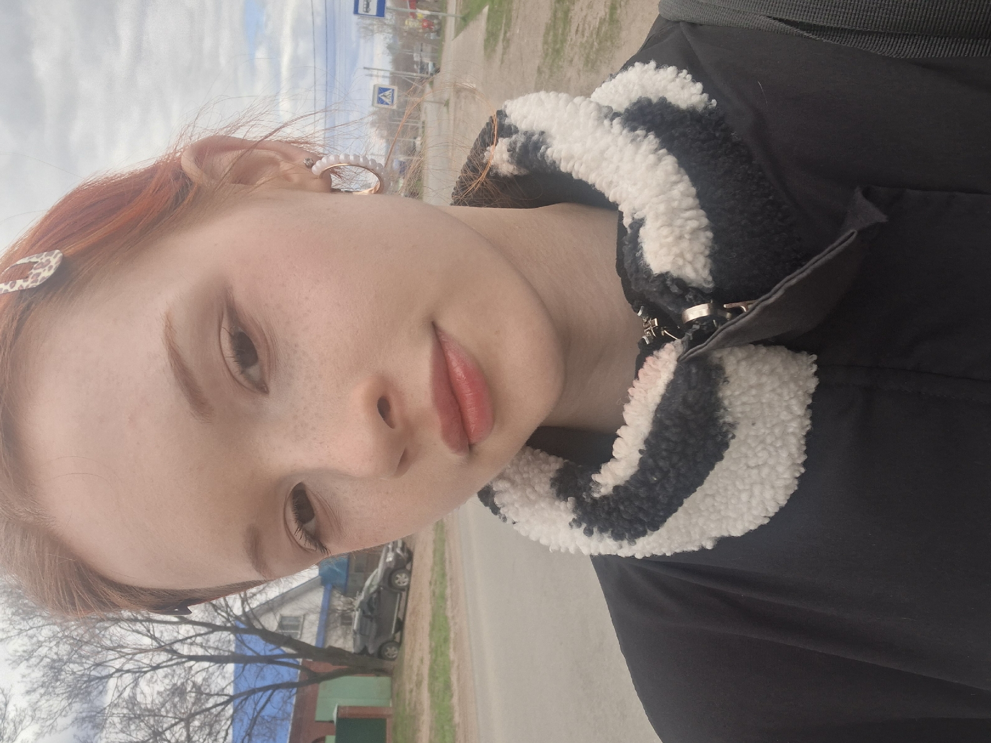 A young woman with reddish-brown hair and visible freckles is captured in a diagonal, headshot-style outdoor photograph in Chaikovsky, Russia. She looks directly at the camera with a subtle, closed-mouth smile, her lips tinted with a soft pink or coral color. Her right eye is slightly narrowed.
She wears a black jacket featuring a prominent, fluffy black and white striped collar, which appears to be made of sherpa or faux fur. An earring with white pearl-like beads adorns her visible right ear, and a small leopard-print hair clip is placed in her hair near her left temple. Her hair appears somewhat windswept.
The background shows an outdoor setting, likely a street or sidewalk. A paved road is visible to the right, with patches of green grass alongside it. In the mid-ground, several bare trees suggest the time of year is late winter or early spring. Distant buildings, possibly houses, can be seen beyond the trees. The sky is bright and partly cloudy. In the upper right background, a blue road sign with white pictograms, one resembling a pedestrian crossing symbol, is discernable. The natural lighting indicates it is daytime.