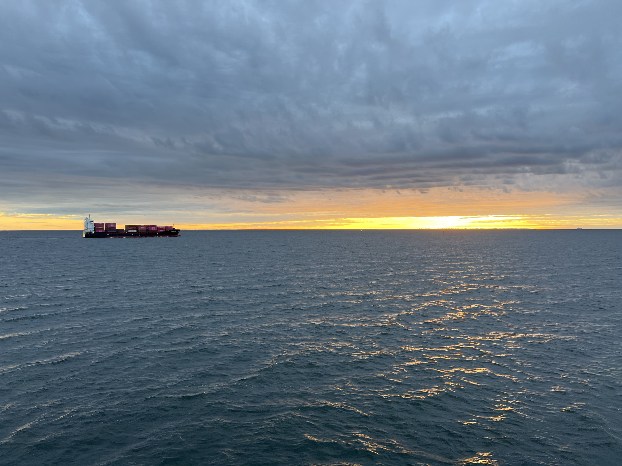The image depicts a cargo ship navigating through calm waters under a dramatic, cloudy sky at sunset. The sky transitions from dark, heavy clouds at the top to a brilliant band of orange and yellow sunlight along the horizon, reflecting on the water's surface. The cargo ship, loaded with stacked containers, is positioned on the left side of the frame, moving away from the viewer. The sea is a deep blue, with gentle ripples that catch the fading light, creating shimmering pathways of gold. There are no people visible in the image. The scene is set on the open sea, with no visible landmass. While the image depicts a maritime scene, there is no specific indication that it is located in Gdynia, Poland.