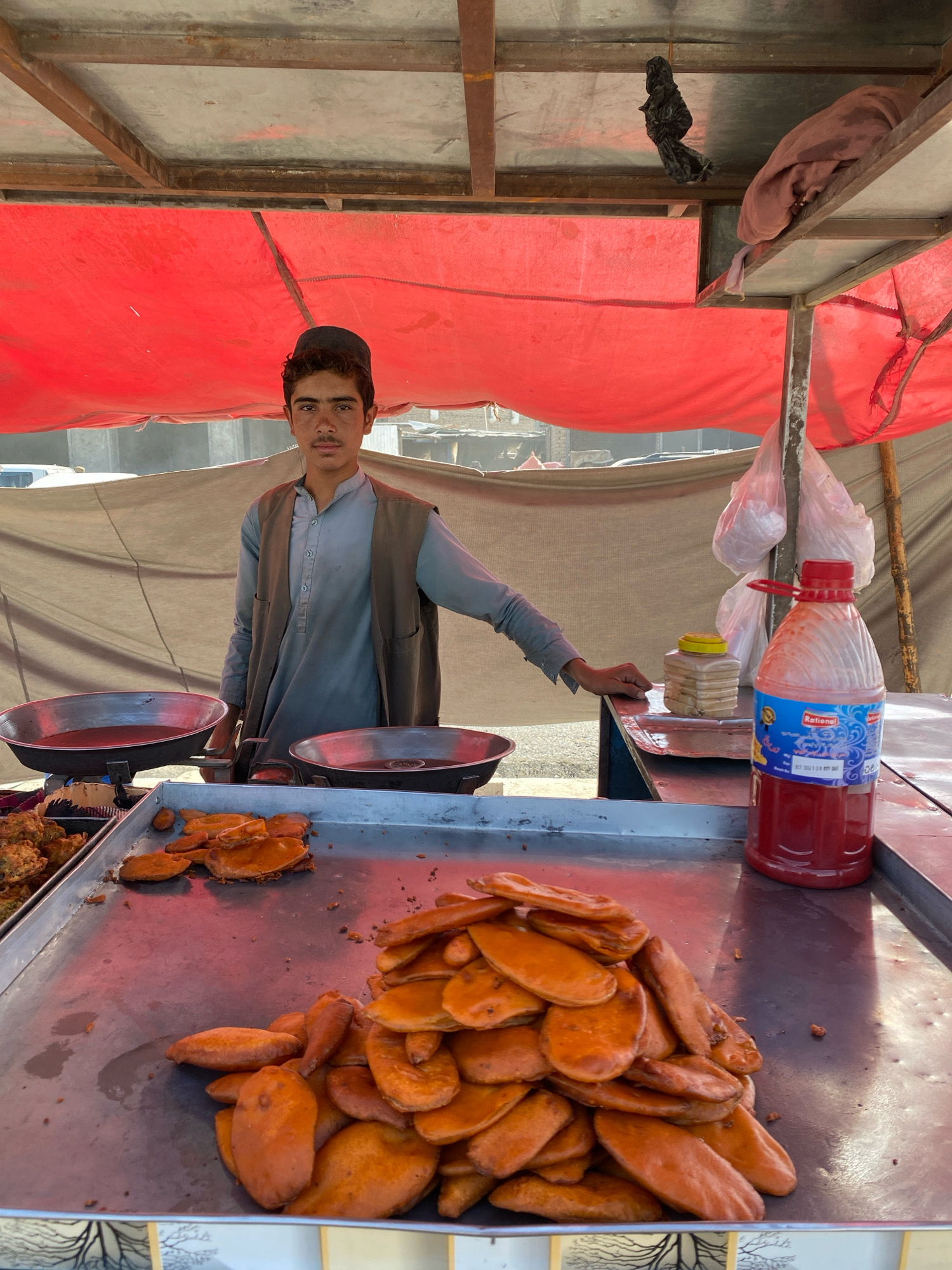 A young man stands behind a food stall in Spin Boldak, Afghanistan. He wears a light blue shirt and a dark vest. In front of him, on a metal tray, are piles of fried pastries. To his left, two large metal bowls contain a dark liquid, possibly oil or sauce. Behind him, a red tarp is draped over a metal frame. A large bottle of red liquid, likely a beverage or condiment, sits on the right side of the stall. The background is blurry, showing hints of buildings and a dusty environment.