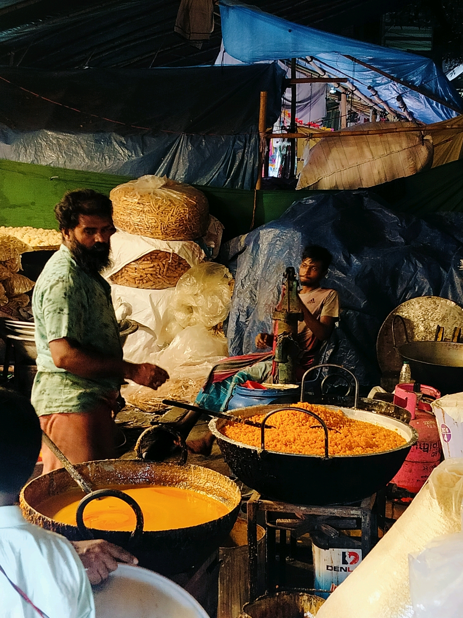 This image depicts a bustling street food stall at night in Dhaka, Bangladesh. A man with a beard, wearing a light green shirt and reddish-brown shorts, stands on the left side of the frame, looking towards the viewer. He appears to be tending to a large, dark pot filled with bright orange food, possibly a sweet or snack. Beside him, stacked high, are large sacks of dry goods, likely snacks or ingredients.
In the background, a younger man, wearing a light-colored t-shirt, operates a manual water pump. Behind him, a large blue tarp is stretched overhead, creating a covered space. The scene is dimly lit, with artificial light sources illuminating the food and the people. Various cooking utensils, pots, and ingredients are visible, suggesting an active food preparation environment. The overall impression is one of a vibrant, albeit somewhat chaotic, nighttime market scene common in Dhaka.