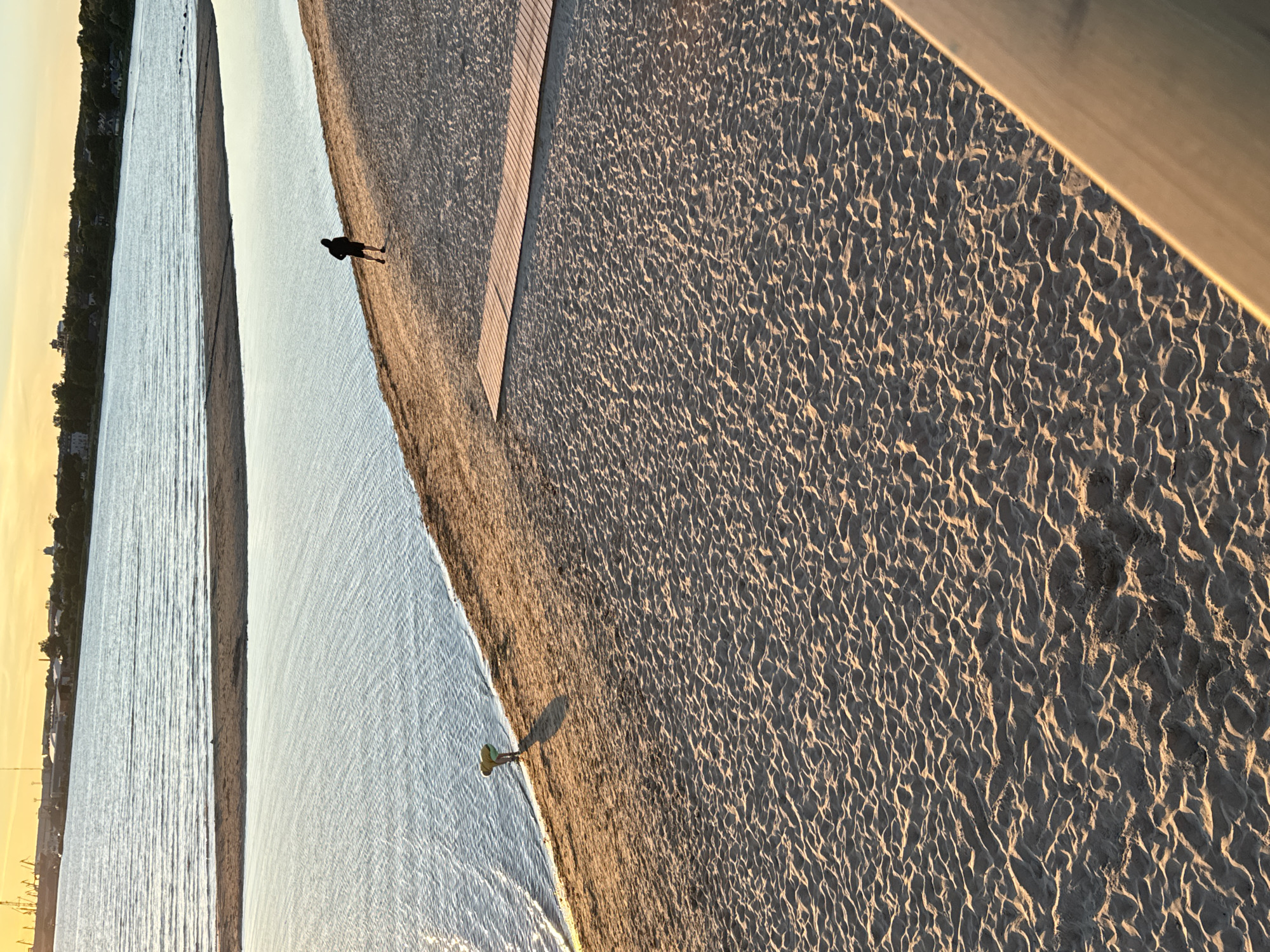 The image depicts a beach scene at sunset, likely in Tallinn, Estonia, given the architectural style of the distant buildings and the presence of industrial elements on the horizon. The foreground is dominated by sandy beach with a wooden boardwalk running diagonally across the sand. In the mid-ground, two individuals are present. One person, dressed in dark clothing, is running along the water's edge. Another person, wearing a yellow top, is bent over at the shoreline, possibly collecting something. The water is calm with gentle ripples reflecting the warm orange and yellow hues of the sunset sky. The background shows a cityscape with a mix of residential buildings, industrial structures, and trees. The overall impression is one of a peaceful evening at a coastal location.