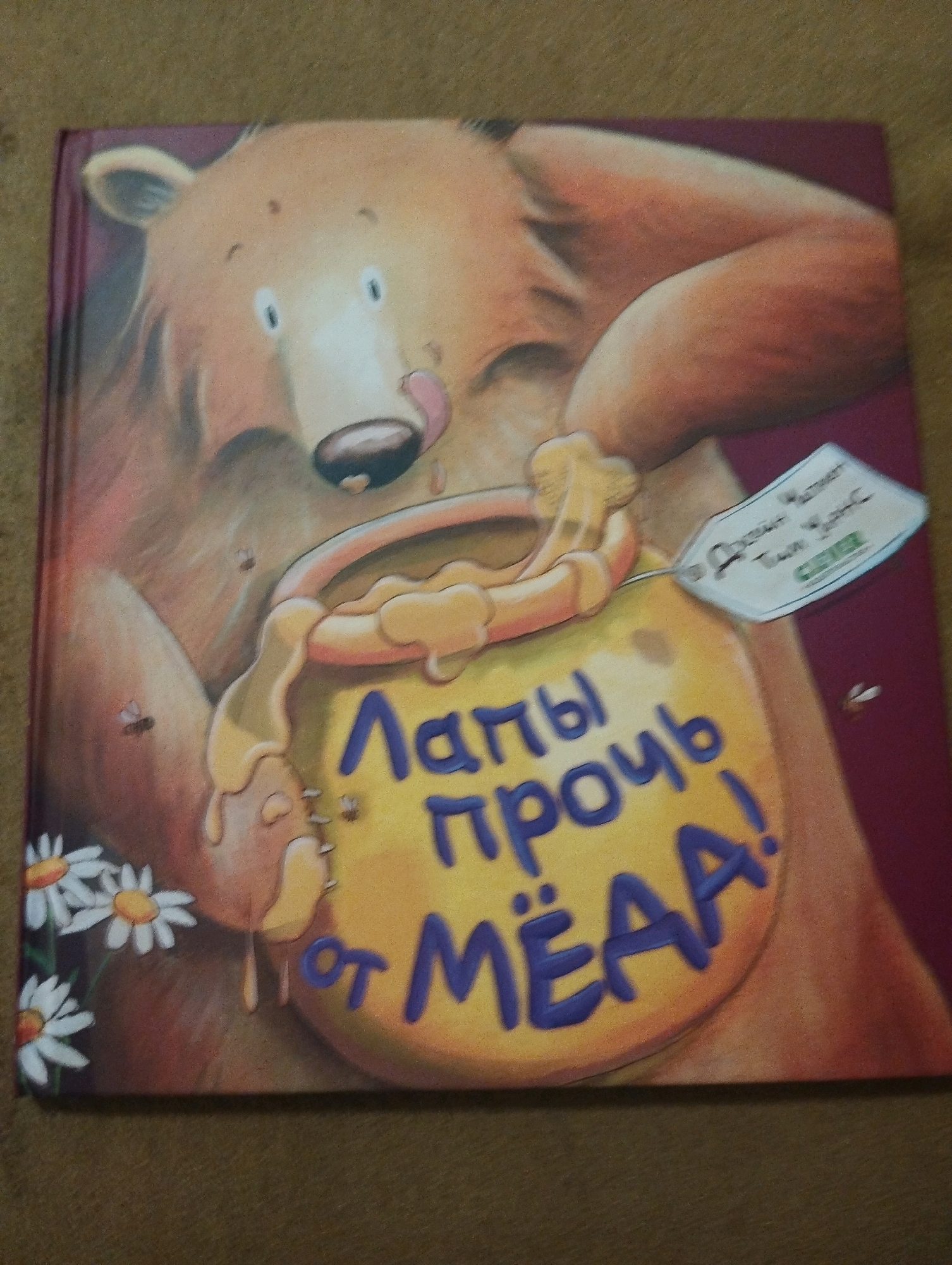 The image shows the cover of a children's book featuring a cartoon bear holding a pot of honey. The bear is depicted with its tongue sticking out, licking the honey. The text on the pot is in Russian and appears to say "Лапы прочь от мёда!" which translates to "Paws off the honey!". There is also a small tag attached to the honey pot with some writing. The background is a plain burgundy color. Daisies are visible on the bottom left of the cover. The book cover is lying on a textured brown surface, possibly a rug or blanket. There is no information in the image that suggests a location in Ashdod, Israel or any other city.