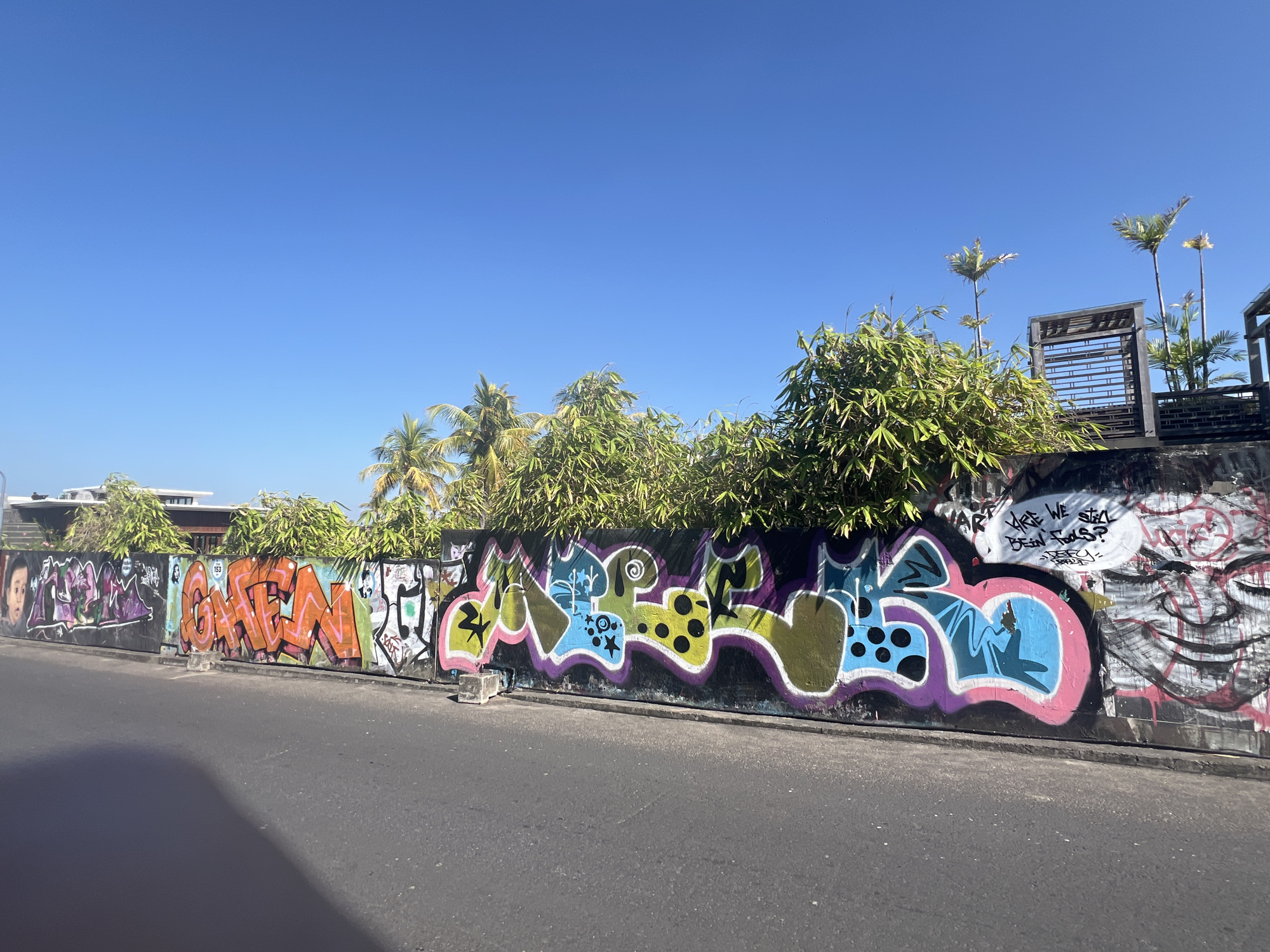 A bright, sunny day is depicted in North Kuta, Indonesia, with a clear blue sky dominating the upper portion of the frame. A long, graffiti-covered wall stretches horizontally across the mid-ground. The wall features a variety of spray-painted artwork, with vibrant colors and diverse styles. Some sections display abstract designs with swirling lines and geometric shapes in shades of purple, pink, green, yellow, and blue. Other parts of the wall are covered in bold, blocky lettering, predominantly in orange and black. There are also depictions of faces and a speech bubble containing text. Lush green foliage, including palm trees and bamboo, grows along the top of the wall, partially obscuring some of the graffiti and creating a natural canopy. In the background, glimpses of buildings and architectural elements, such as wooden structures, are visible above the foliage. A paved road runs along the bottom of the image, casting a shadow onto the asphalt. The scene suggests an urban environment decorated with street art, typical of a tropical setting.