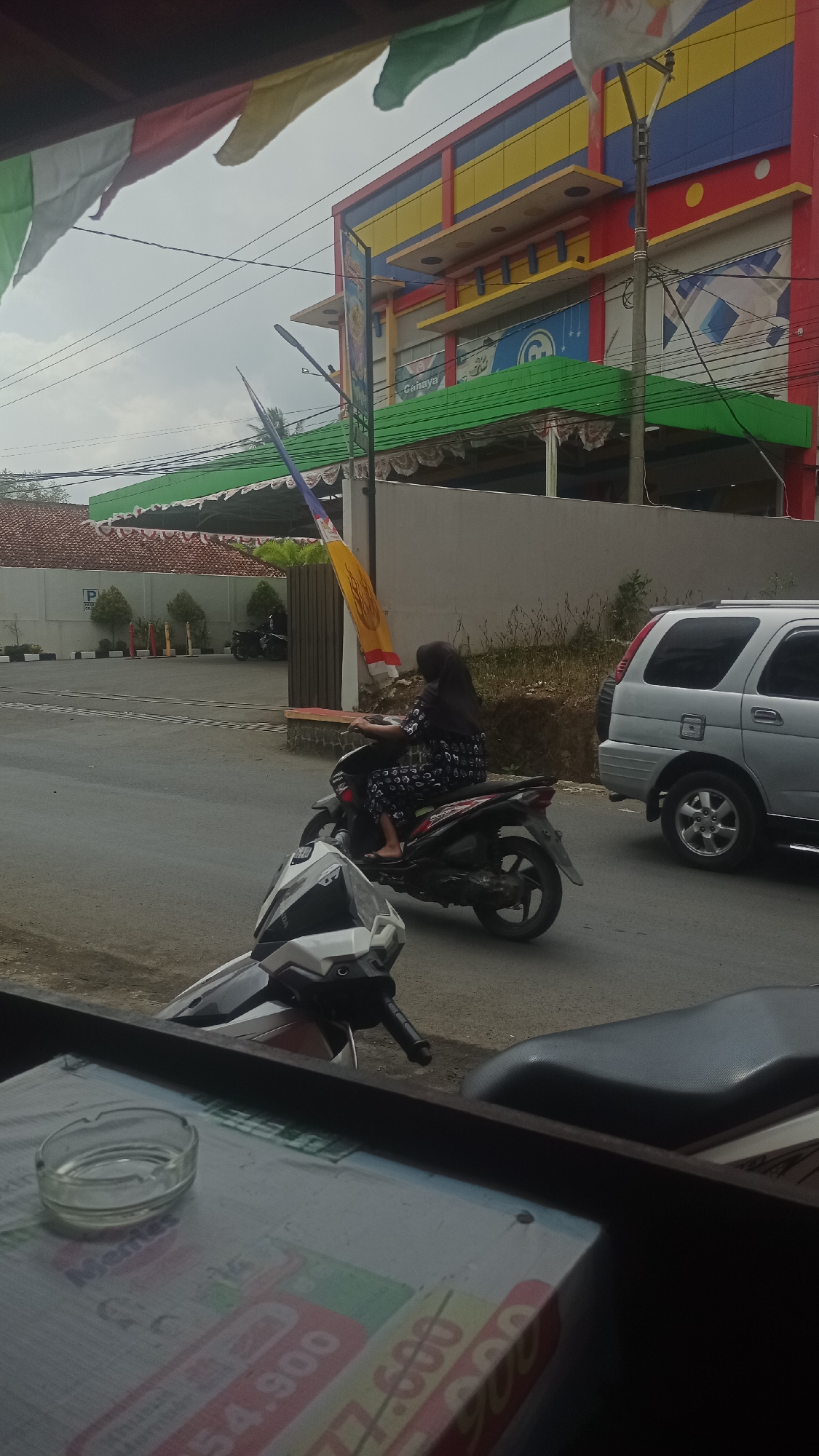 A woman wearing a black hijab and a patterned dress rides a dark-colored motorcycle on a paved road. She is positioned in the center of the frame, moving from left to right. To her right, a silver SUV is also present on the road. In the background, a multi-story building with colorful red, yellow, and blue accents stands. Green awnings extend from the building, and several flags are visible. The scene appears to be located in an urban or semi-urban area. The foreground shows a portion of a table with an ashtray and some printed material. The sky is overcast.