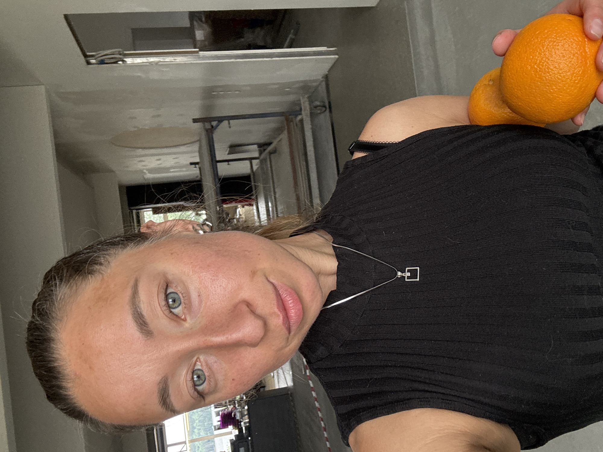A woman with light skin, blue eyes, and dark hair is holding two oranges near her shoulder. She is wearing a black ribbed top and a silver necklace with a square pendant. The background shows an interior space with exposed ceilings and visible construction or renovation. The location is Chaikovsky, Russia, based on contextual information not explicitly visible in the image.