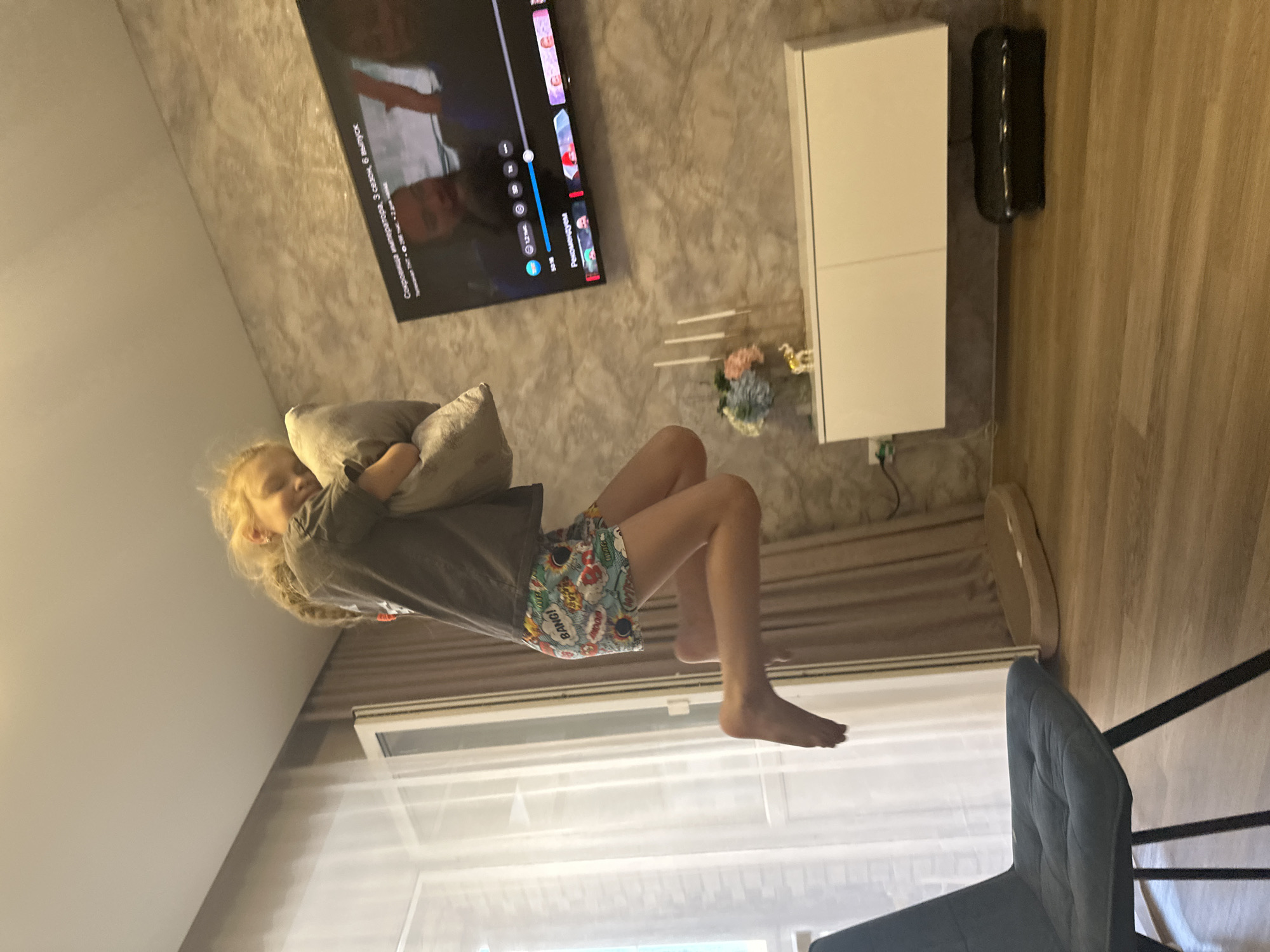 A young blonde girl in a grey t-shirt and colorful comic-themed shorts is captured mid-air, appearing to jump while hugging a light grey pillow. She is positioned in the center of a room with wood-paneled flooring. To her left, a dark grey upholstered chair with black metal legs is visible. Further left, a sheer white curtain hangs in front of a window, with vertical blinds. Behind the girl, a textured grey wall serves as a backdrop. A flat-screen television is mounted on this wall, displaying a scene with two individuals inside a vehicle, and text in Russian below it. To the right of the television, a white cabinet is mounted on the wall, with a decorative floral arrangement and small elephant figurine on top. The overall environment suggests an indoor residential setting.