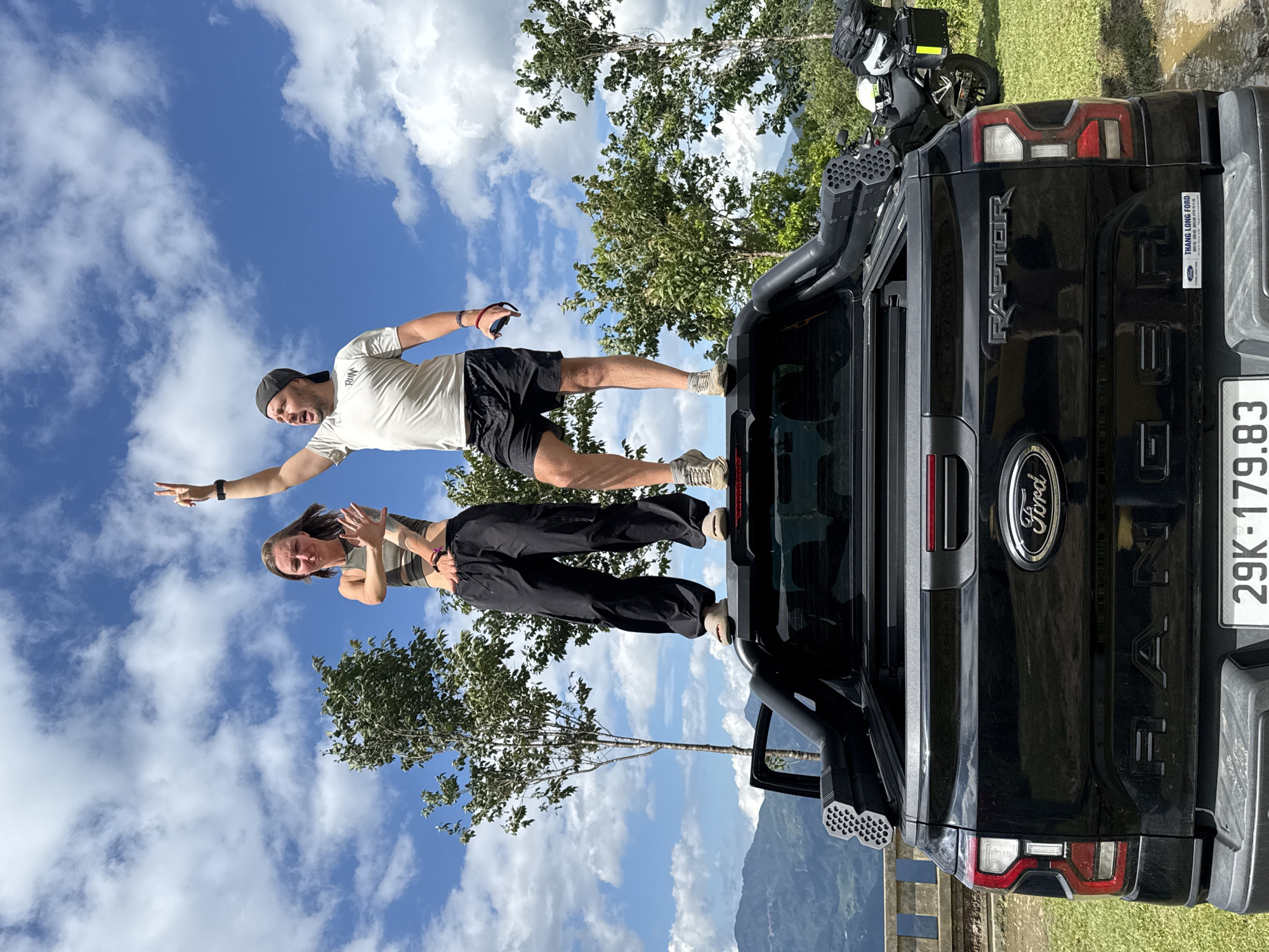 Two individuals, a female and a male, are standing on the bed of a black Ford Ranger Raptor pickup truck, positioned against a clear blue sky with scattered white clouds. The scene is set in a mountainous outdoor environment, identified as Tu Le, Vietnam.
The female, on the left, wears a grey-green sports bra and black cargo pants. Her dark hair is loose, and she has a direct expression, extending both hands forward with fingers spread. She wears a dark bracelet on her left wrist and light-colored slip-on shoes.
The male, on the right, wears a white short-sleeved t-shirt with "RUN" printed on the chest, black shorts, and a grey baseball cap worn backward. He has a beard and is actively gesturing with his right arm raised, forming a V-sign with his fingers (peace sign). His left hand holds a dark object, possibly a phone or camera remote. He wears a dark smartwatch on his left wrist, a red and blue bracelet on his right wrist, and light-colored athletic shoes.
The black Ford Ranger Raptor pickup truck is prominent in the foreground. The Ford logo is visible on the tailgate, along with "RAPTOR" embossed above it and "RANGER" embossed below. A sticker for "THANG LONG FORD" with a phone number is affixed to the right side of the tailgate. The license plate, "29K-179.83," is clearly visible. The truck bed is equipped with a black roll-top tonneau cover and a black bed rack system featuring what appears to be a multi-chambered snorkel or air intake system on the left side.
In the background, behind the truck and to the right, a black adventure motorcycle with saddlebags and two helmets (one white, one yellow) is parked on a grassy patch. Further back, green trees are scattered across the landscape, and a series of verdant mountains rise under the bright sky. A stone railing or barrier is partially visible behind the truck on the left, suggesting a viewpoint or elevated area. The ground where the motorcycle is parked appears to be a mix of grass and dirt.