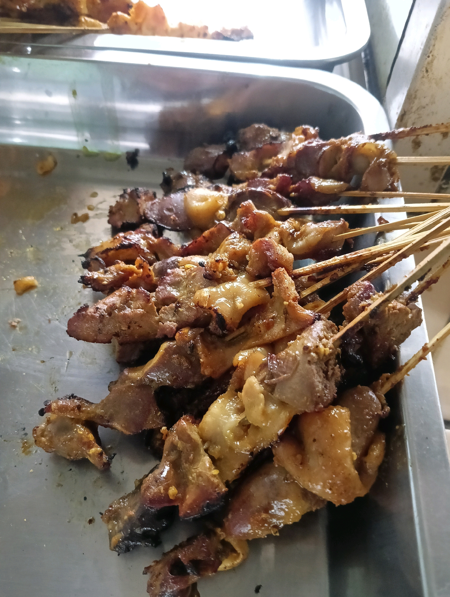 A close-up, high-angle shot shows a metal tray filled with numerous skewers of grilled meat. The meat appears to be chicken or other poultry, with some pieces exhibiting slightly charred edges. The skewers are made of thin, light-colored wood and are arranged in a dense cluster, obscuring much of the tray beneath. The background is blurred but suggests an indoor setting, possibly a food stall or restaurant in Sokaraja Kidul, Indonesia, given the context. No people are visible in the frame.