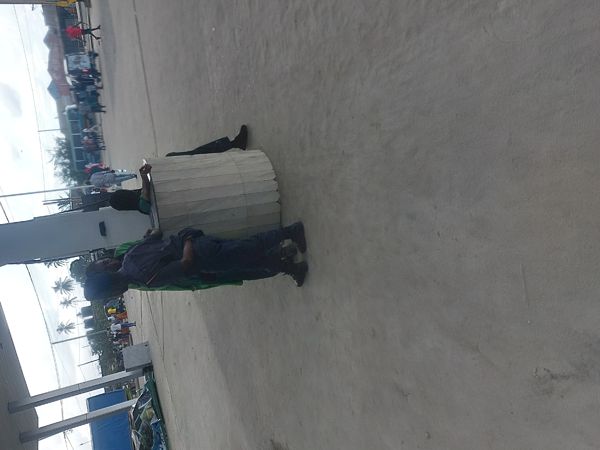 A long-angle shot depicts a location in Dar es-Salaam, Tanzania. A man is lying on his side, resting his head on a cylindrical, beige object composed of vertically oriented sections. He is wearing a dark blue uniform, possibly security or official attire. Behind him, another person is lying flat, partially obscured by the cylindrical object, with only their legs visible. A line of pedestrians and buildings can be seen in the background. In the foreground, there is a partial view of a shaded area with blue tarps. The setting appears to be a public space or square.