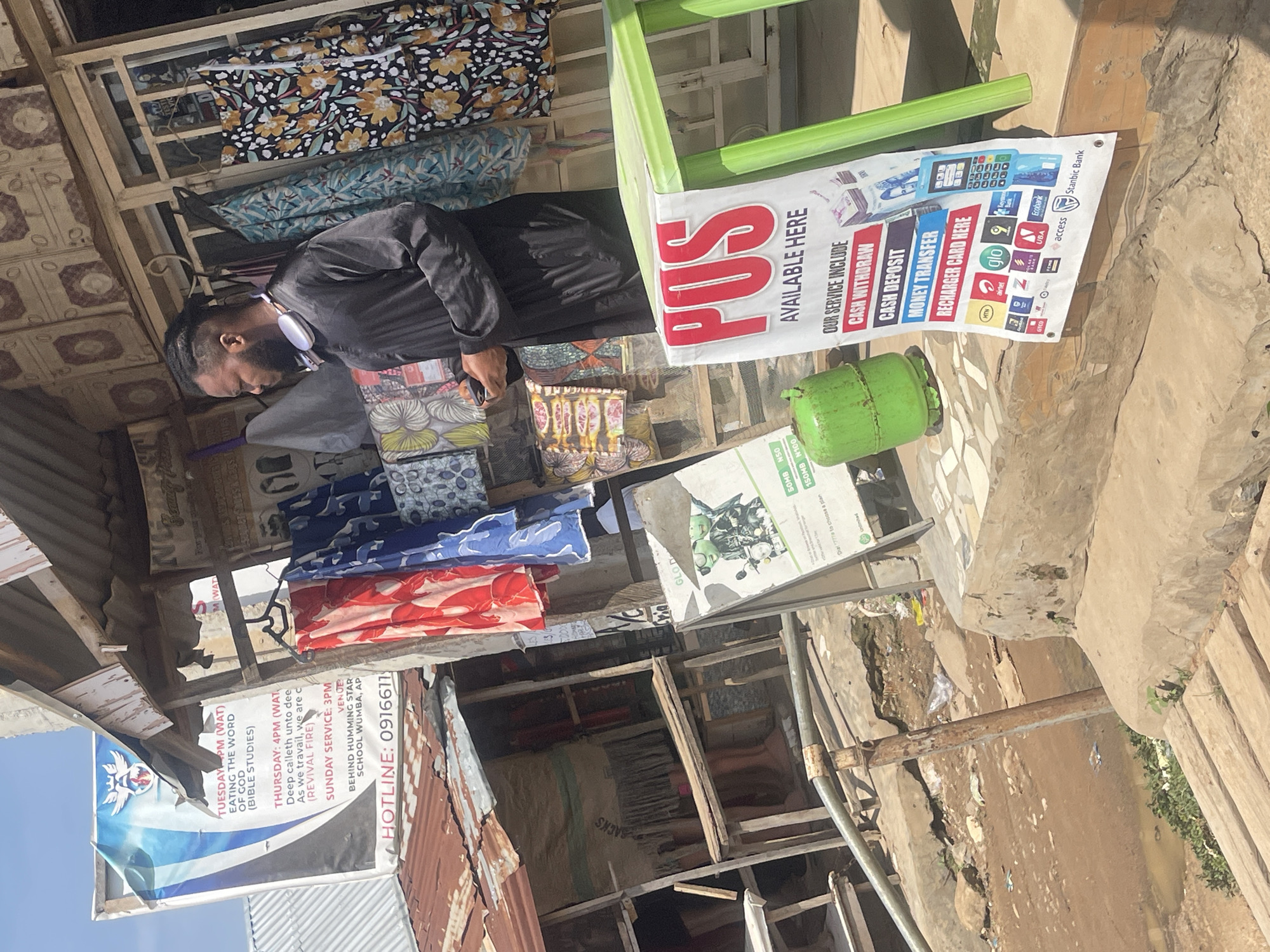 In an outdoor market stall in Abuja, Nigeria, a young man stands looking down at his phone. He is wearing a dark, flowing garment and white headphones around his neck. The stall is filled with colorful fabrics, and a sign advertising "POS Available Here" is prominently displayed on a green table. Behind the man, a poster for a church service lists a "HOTLINE: 0916611..." The ground is dirt and appears uneven, with some puddles of water. The stall appears to be made of corrugated metal and wood, with some signs of age and wear. Other objects visible include a green gas cylinder, a metal railing, and some mannequins in the background. The scene suggests a bustling commercial area.