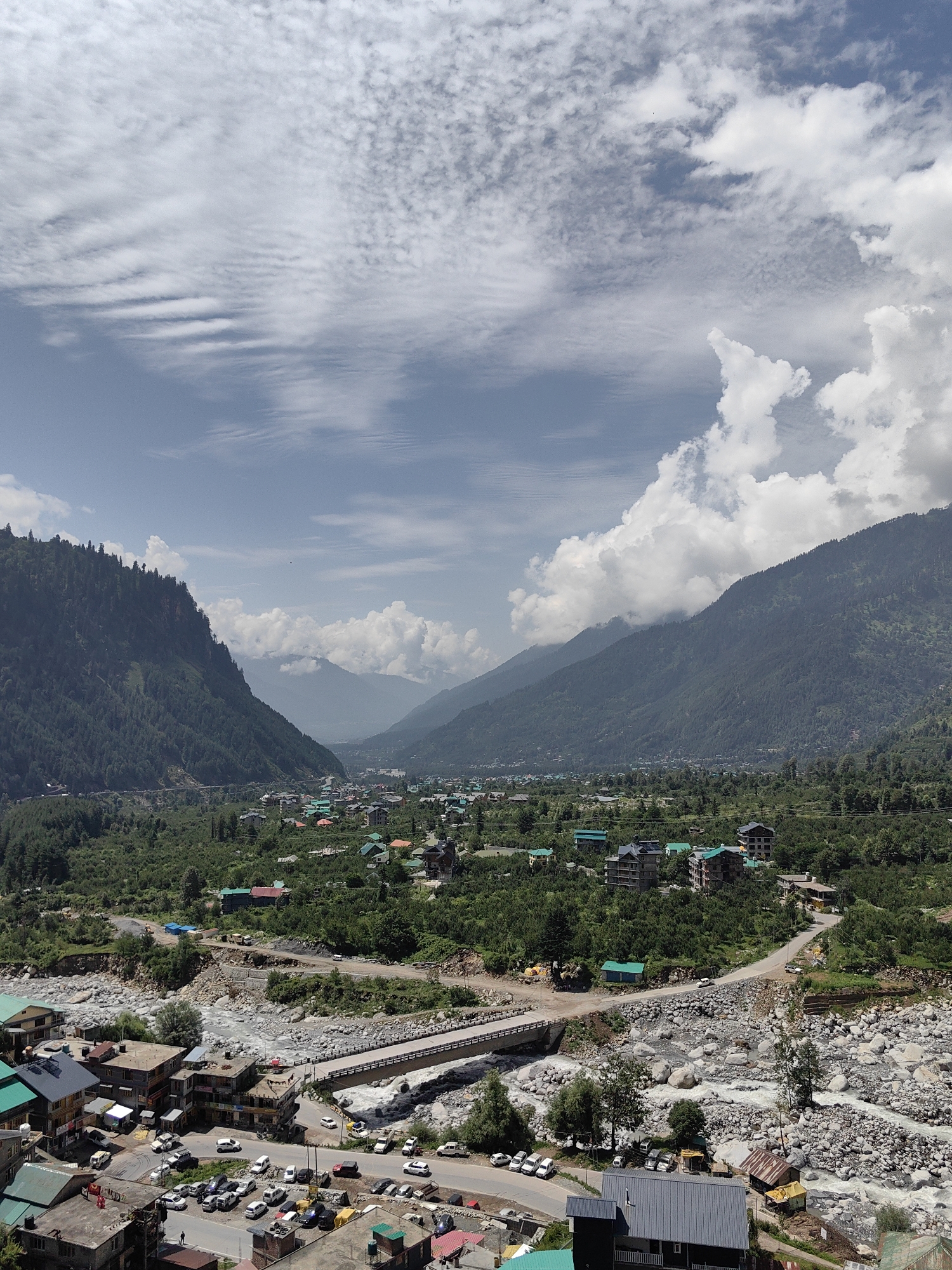 The image captures a wide-angle view of a valley in the city of Kullu, India, dominated by lush green mountains and a meandering river. In the foreground, a cluster of buildings with multi-colored roofs, characteristic of the region, lines a road packed with parked cars. A concrete bridge spans the rocky riverbed, connecting different parts of the settlement. Beyond the immediate vicinity, the valley opens up to reveal more buildings nestled amidst dense vegetation, stretching towards the majestic, tree-covered slopes of the mountains. The sky above is a canvas of white, fluffy clouds against a pale blue expanse, with some areas showing dappled sunlight, indicating a partly cloudy day. The overall scene depicts a serene, mountainous landscape coexisting with a small, vibrant community.