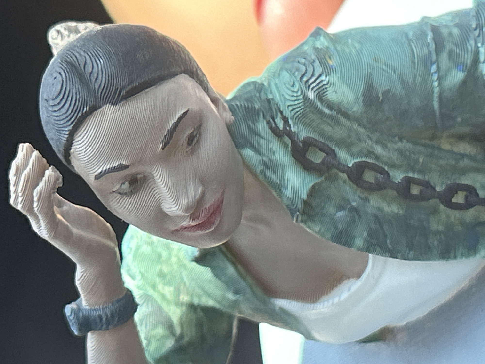 The image is a close-up, high-angle shot of a 3D-printed figurine of a woman. The figurine is depicted from the chest up, with her head tilted downwards and to the left, and her right arm raised towards her head. She is wearing a green, patterned jacket over a white tank top. The jacket has a black chain design across the front. She has dark hair pulled back and a dark bracelet on her left wrist. The texture of the 3D printing process is visible, characterized by visible layer lines throughout the figurine. The background is blurred and appears to be a mix of dark and orange tones, suggesting an indoor setting. The location in the city of Lozen, Bulgaria, is not discernible from the image.