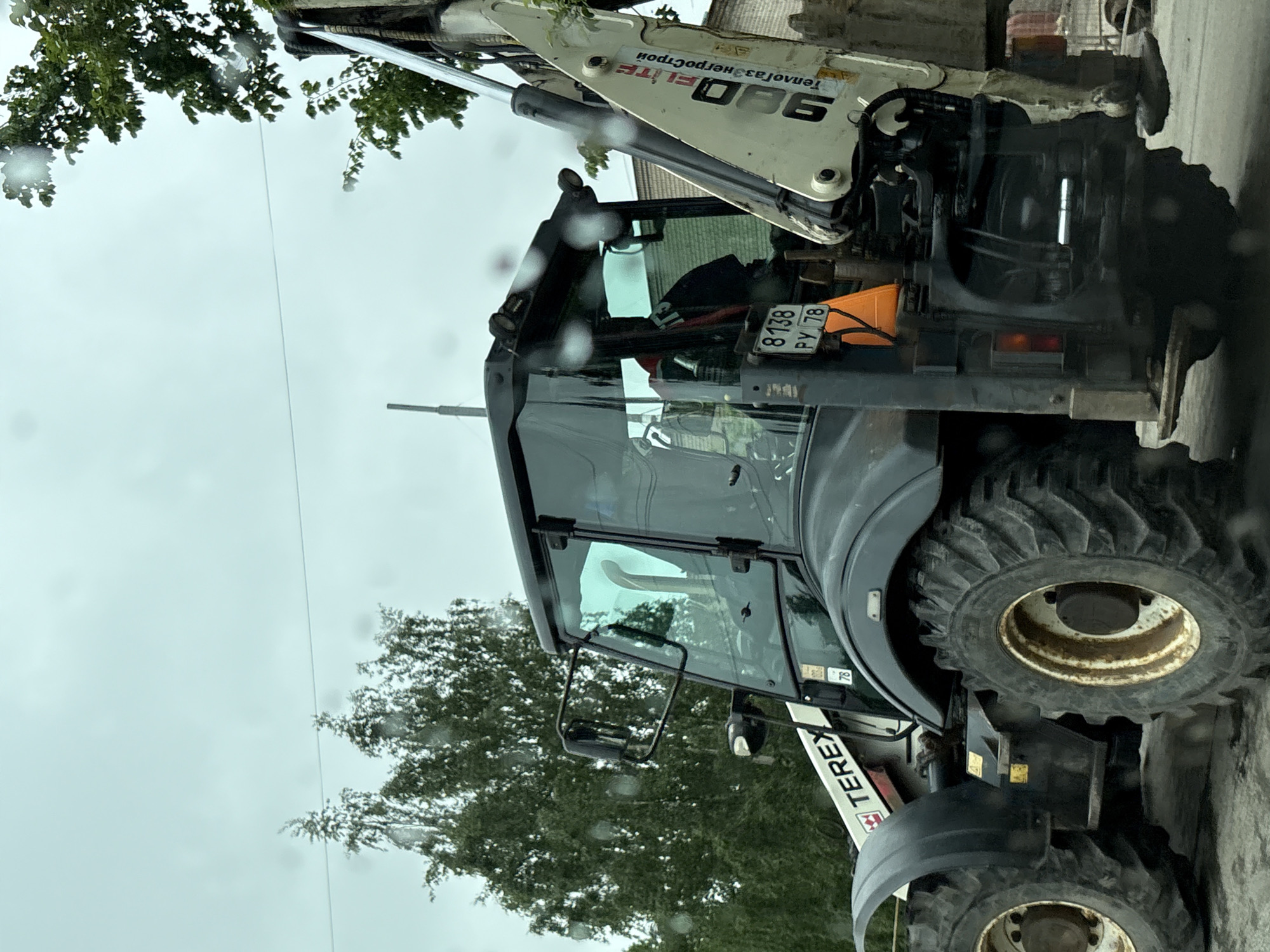 The image shows a Terex 960 Elite backhoe loader with the license plate 8138 PY 78, indicating the vehicle is registered in Saint Petersburg, Russia. The backhoe loader is parked on a street, likely within the city. The weather appears overcast. Trees are visible in the background. The backhoe loader's arm is extended upward.