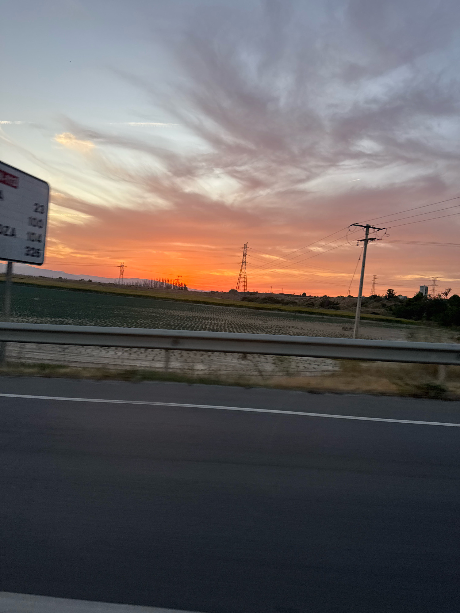 This is a vertical, eye-level, medium shot of a sunset over a rural landscape in Valtierra, Spain, as viewed from a moving vehicle on a highway. The sky is filled with an orange and pink gradient due to the sunset, with wispy clouds scattered across it. In the foreground, the dark asphalt of the highway is visible, with a white line running parallel to it. A metal guardrail separates the highway from a field of crops, which are a deep green. Beyond the field, several power lines and transmission towers are visible, along with a faint line of mountains in the distance. A road sign is partially visible on the left side of the image, displaying numbers and text. The overall scene captures a moment of natural beauty during sunset in an agricultural region.
