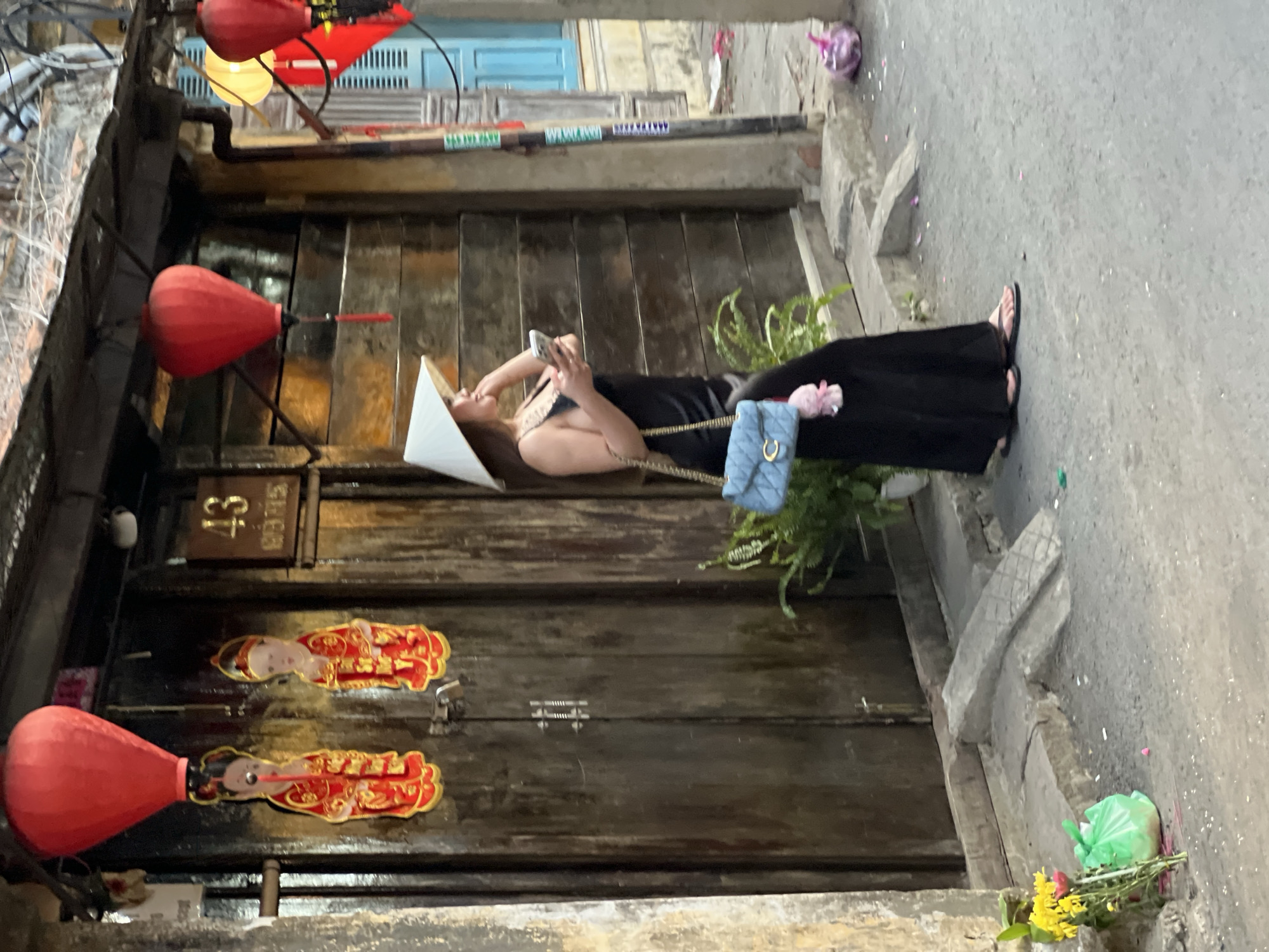 A woman wearing a conical hat and a black dress is standing on a street in Hoi An Ancient Town, Vietnam. She is holding a smartphone in her right hand and a light blue quilted handbag over her left shoulder. The handbag has a C-shaped clasp and a pink charm hanging from it. Behind her is a weathered wooden door with two red figures with gold trim hanging on it. Above the door, there is a red paper lantern. To the right of the door, a wooden plank wall is visible, and a blue-shuttered window is further to the right. In front of the door, there is a potted fern. On the ground to the left, there is a bunch of yellow and pink flowers with a green plastic bag. The street is paved with asphalt and has a concrete curb. In the background, there are more buildings and a light yellow lantern.