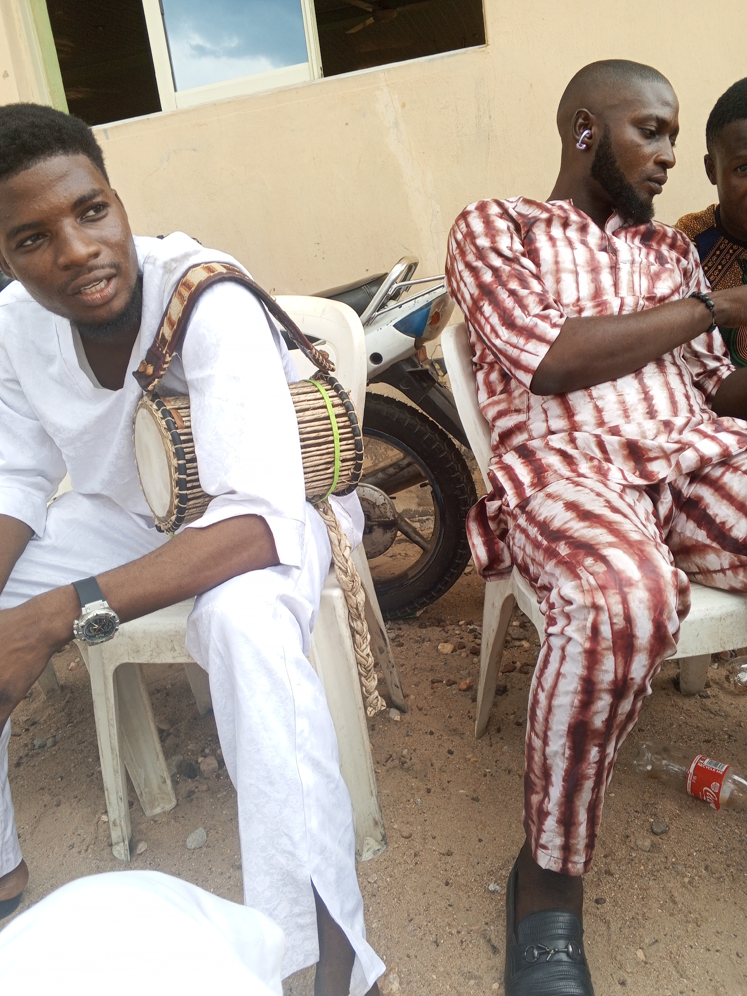 The image shows two men seated on white plastic chairs in an outdoor environment in Zango-ndaji, Nigeria. The man on the left wears a white traditional outfit and a watch, and a small drum hangs on his shoulder. The man on the right is dressed in a red and white tie-dye patterned suit and black shoes, and he sports an earring. A motorcycle is parked behind them. A Coca-Cola bottle is on the ground near the man on the right. The ground appears to be sandy.