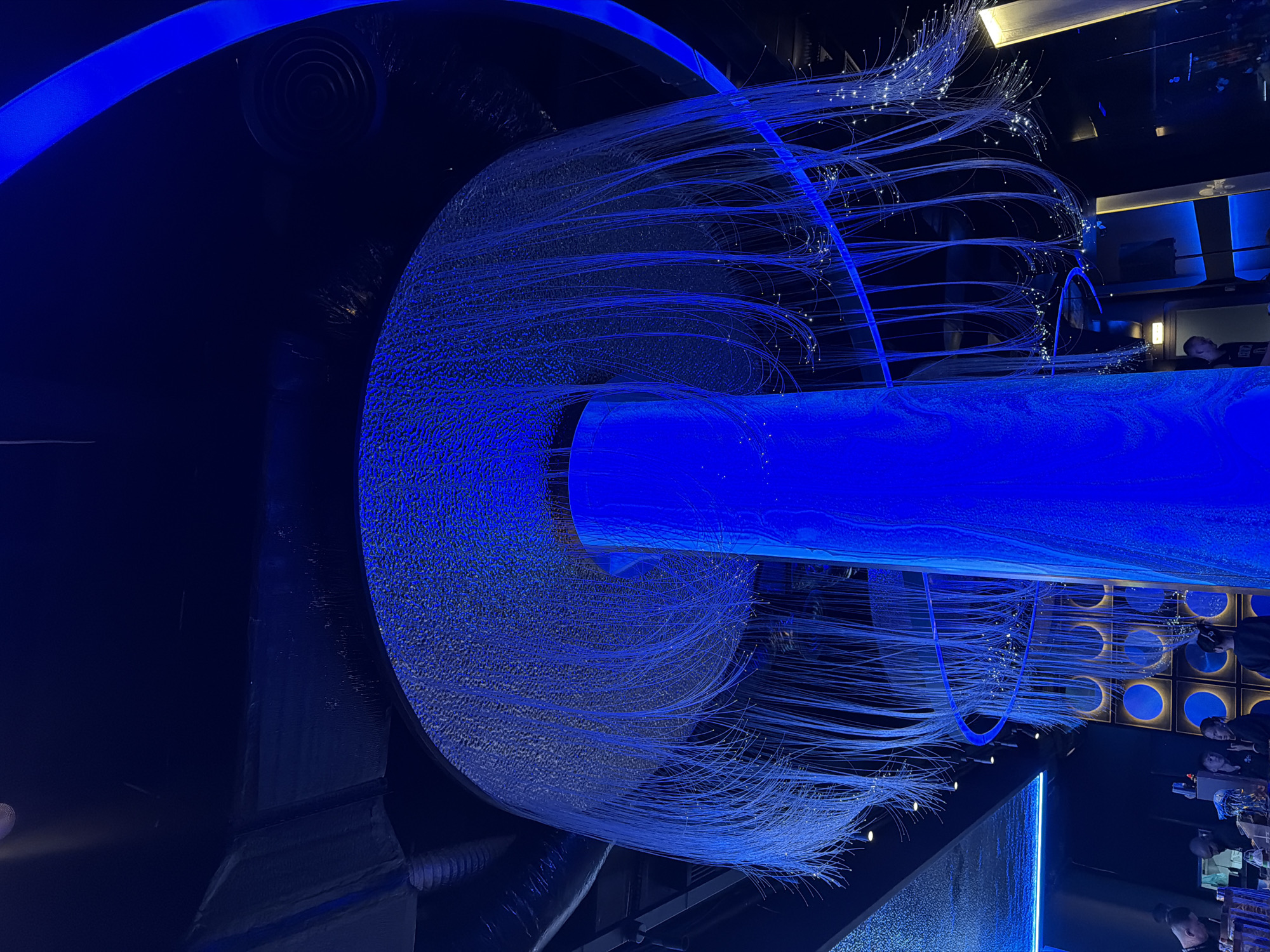 The image depicts the interior of a dimly lit establishment, likely a restaurant or bar, in Nha Trang Ward, Vietnam. Dominating the scene are two large, cylindrical structures covered in what appears to be a vibrant blue light. These structures are adorned with numerous thin, fiber-optic strands that cascade downwards, some ending in small, glowing points of light. Above, a large, circular canopy, also illuminated with blue light and speckled with a textured pattern, is suspended. A thin, curved blue band of light encircles the upper portion of the space. In the background, several individuals are visible, appearing to be seated at tables and interacting with each other. The ambiance is modern and futuristic, characterized by the extensive use of blue lighting and sleek design elements.