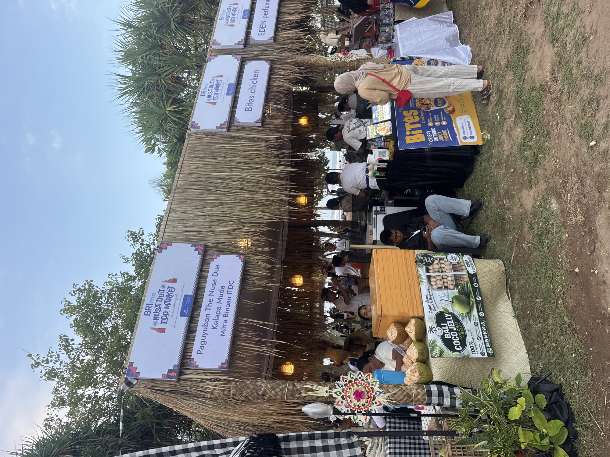 An outdoor market stall in Kuta Selatan, Indonesia, features a thatched roof structure displaying various signs for vendors. One sign reads "BRImo NUSA DUA ECO MARKET," and another advertises "Paguyuban The Nusa Dua Kelapa Muda Mitra Binaan ITDC." Other signs indicate "Bites chicken" and "EDEN perfume." The stall sells products including coconuts and what appears to be "BALI COCO JELLY." Several people are present, including a woman in a headscarf looking at a phone, a man sitting and using his phone, and vendors attending to customers. The background shows palm trees and a beach with the ocean visible. The overall atmosphere is that of a bustling marketplace on a tropical coast.