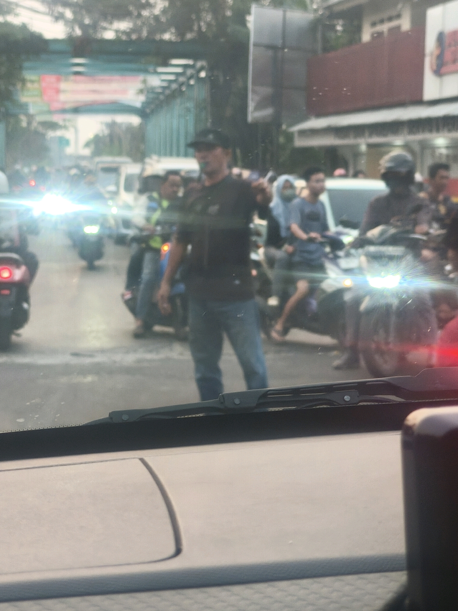 The image shows a street scene in Karangsatria, Indonesia, likely during daylight hours. The foreground is dominated by the interior of a vehicle, with the windshield and part of the dashboard visible, suggesting the photo was taken from a driver or passenger's perspective. Outside, a busy street is depicted with multiple motorcycles and a few pedestrians.
In the center of the frame, a man wearing a dark t-shirt, dark pants, and a black baseball cap is standing and appears to be directing traffic. He is holding his right arm up, with his palm facing forward, in a gesture that indicates stopping or directing. Several motorcycles are on the road, some with riders. One motorcycle in the foreground has its headlights illuminated brightly. The environment appears to be a typical urban or suburban street with buildings and trees lining the sides. The overall impression is of a traffic intersection or a busy roadway with ongoing activity.