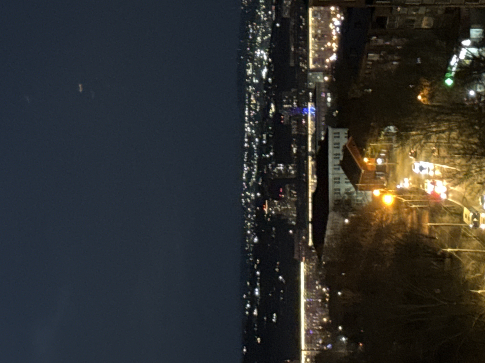 The image shows a night view of Krasnoyarsk, Russia. The sky is dark, transitioning to a brightly lit cityscape with numerous buildings and streetlights. Visible are a mix of residential buildings, illuminated roads with moving vehicle lights, and dense vegetation, probably trees.