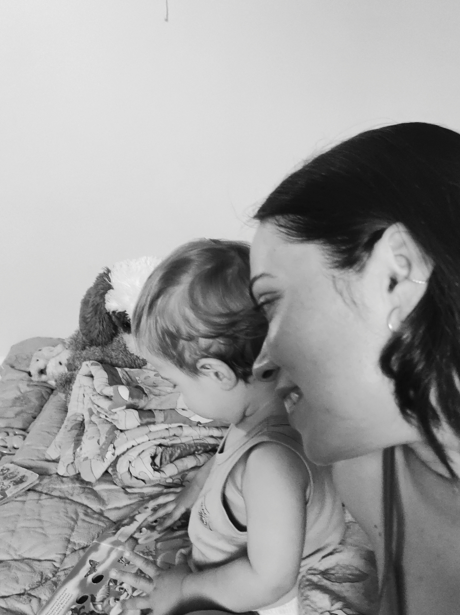 The black and white photograph shows a woman and a young child sitting on a bed in what appears to be a private residence in Unknown, Unknown. The woman, positioned on the right side of the frame, has dark hair and is looking towards the left. A small earring is visible in her left ear. A young child with light hair is sitting next to her, also facing left. The child is wearing a tank top and appears to be holding or looking at a book or object with holes in it. The bed is covered with a patterned blanket or quilt. To the left of the child, a stuffed animal can be seen on the bed. The background is a plain white wall.