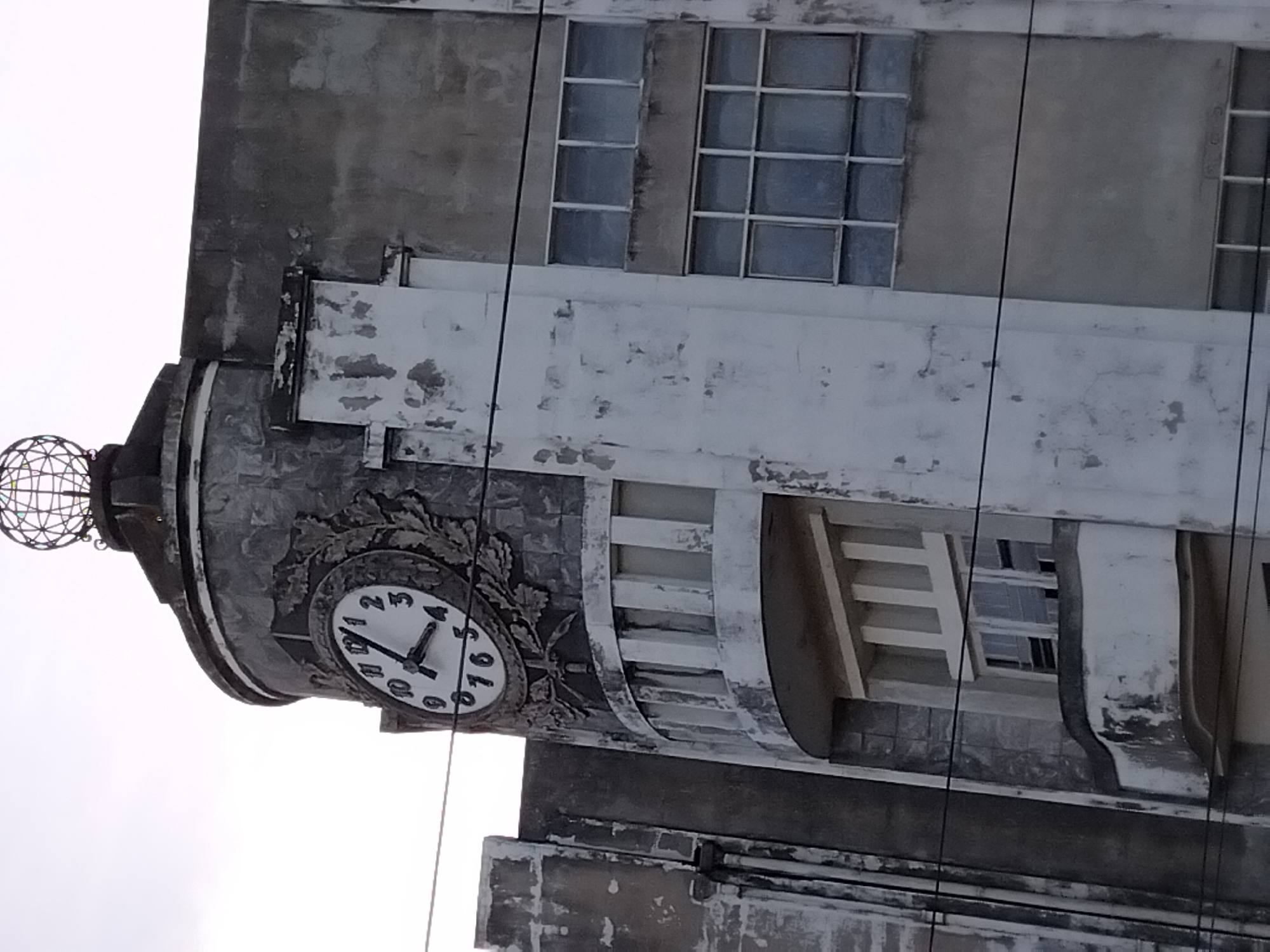 The image shows a close-up, low-angle exterior view of the clock tower and facade of the former Estação de Camionagem in Vila Nova de Famalicão, Portugal. The clock face, adorned with a wreath, is visible on the cylindrical section of the tower. The building exhibits an art deco architectural style, characterized by geometric shapes and decorative elements. The facade is a mix of concrete and what appears to be tiled sections, showing signs of age and weathering with peeling paint and discoloration. Several windows of varying sizes and designs are visible on the building. Overhead, power lines cross the frame. The sky is overcast. No people are present in the image.