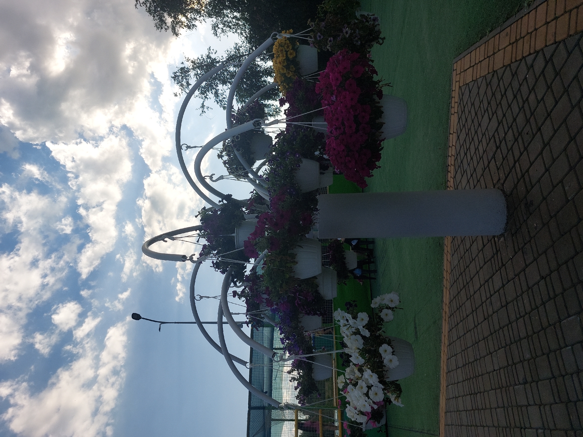 An outdoor, eye-level, full shot captures a decorative floral display against a sky with scattered clouds. The floral display is a tiered arrangement of white, pink, purple, and yellow petunias hanging in white pots from a white, arched metal structure. The structure is supported by a thick, white central column. The ground beneath the display is paved with interlocking rectangular bricks, with a strip of yellow bricks bordering a green artificial turf area. In the background, out of focus, a tall, black lamppost is visible against the sky. There are also glimpses of trees and a fence. No people are clearly visible or identifiable. The overall scene suggests a public garden or park setting.