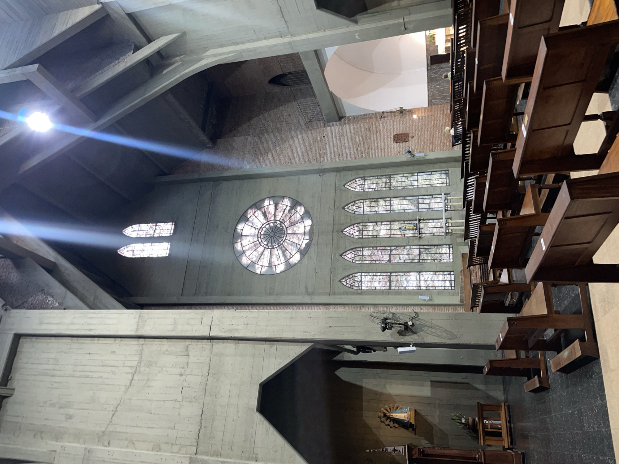 This image captures the interior of a church, characterized by its modern and robust architecture, likely in Vicente López, Argentina. The perspective looks across a vast space with high ceilings.
The structure features prominent, exposed concrete beams and walls, alongside sections of reddish-brown brickwork, contributing to a substantial, almost brutalist aesthetic. On the right side of the frame, rows of dark wooden pews are arranged, indicating seating for a congregation.
A large, circular rose window with intricate stained glass patterns dominates a side wall, allowing bright natural light to flood the interior and creating noticeable lens flares. To its right, a series of tall, arched stained glass windows are vertically aligned, further illuminating the space. Another smaller, arched window is visible higher up.
Near the bottom right, a small devotional area is set up, featuring a framed religious image, possibly of the Virgin Mary, and some floral arrangements. Another piece of religious art is mounted on a concrete wall section nearby. Ceiling fans and various light fixtures are attached to the structural elements. High up in a brick wall section, a metal railing or ladder leads to an opening.
A few individuals are visible: one person is partially discernible kneeling or sitting in the lower right, and others can be faintly seen among the pews, suggesting a quiet moment of prayer or contemplation. The bright natural light indicates it is daytime. On a brick wall, a plaque with stylized script is visible, though the text is unreadable. The overall atmosphere conveys solemnity and architectural grandeur.