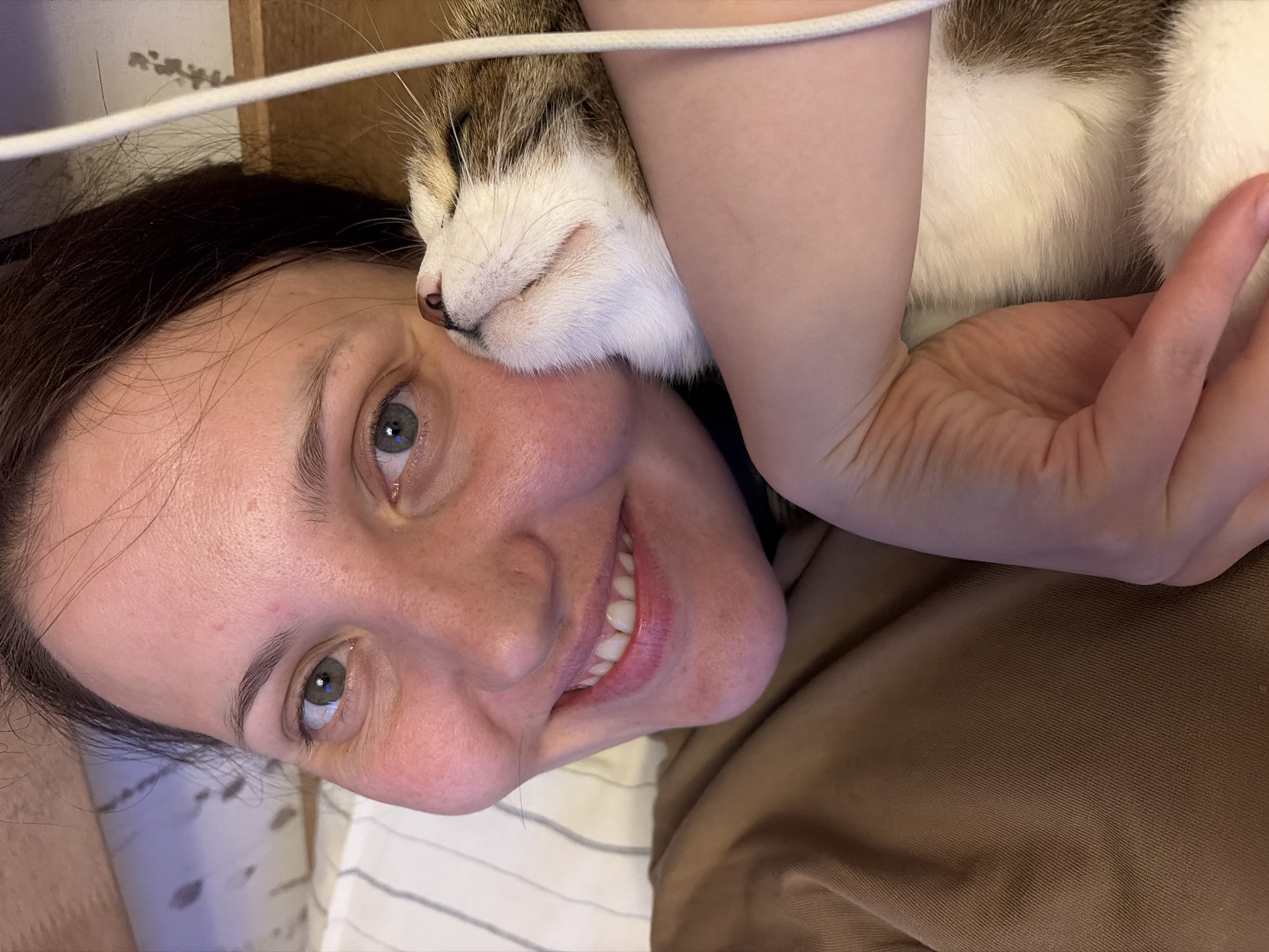 An indoor close-up image depicts an adult female and a domestic cat in close proximity. The female, positioned on the left, has light skin, dark hair, and green eyes, and is smiling with visible teeth. She is wearing a brown-colored garment. A domestic cat, displaying white fur with brown and tan tabby markings, is situated on the right side of the frame, with its eyes closed and its face pressed against the female's right cheek. A light-skinned arm, likely belonging to the female, is visible in the lower right, seemingly embracing the cat. A white cord runs vertically along the right edge, partially obscuring the cat's head. The background includes a striped light-colored fabric on the upper left and a dark wooden surface on the upper right. The specific location within the city of Chaikovsky, Russia, is not discernible from the provided image.