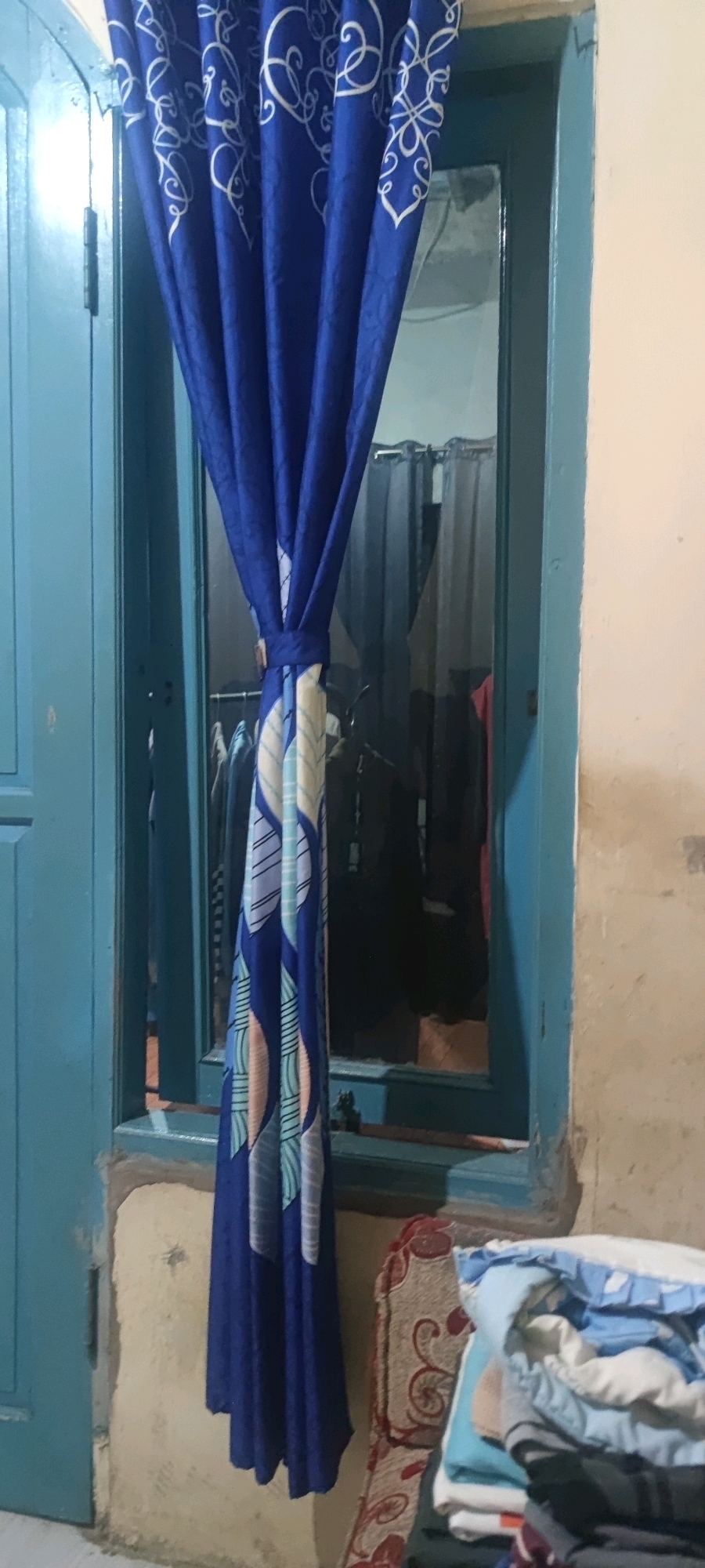 A medium-shot image shows a blue curtain with a white swirling pattern and a leaf-like pattern in blue, white, and pinkish hues, tied back by a matching fabric band. The curtain hangs on the left side of a blue-framed window, partially obscuring the view. Through the window, several dark garments are visible hanging on a rack, along with sheer grey curtains in the background.
On the right side of the image, a stack of folded clothing, predominantly in white and blue, is placed on a patterned fabric surface that appears to be a rug or upholstery. The wall on the right is painted a light, textured color. The bottom of the window frame shows some wear and tear. The overall scene suggests an interior space, possibly a room or closet, with an emphasis on textiles. The image is taken indoors, with the lighting appearing to be artificial. The location is identified as Gambarsari, Indonesia.