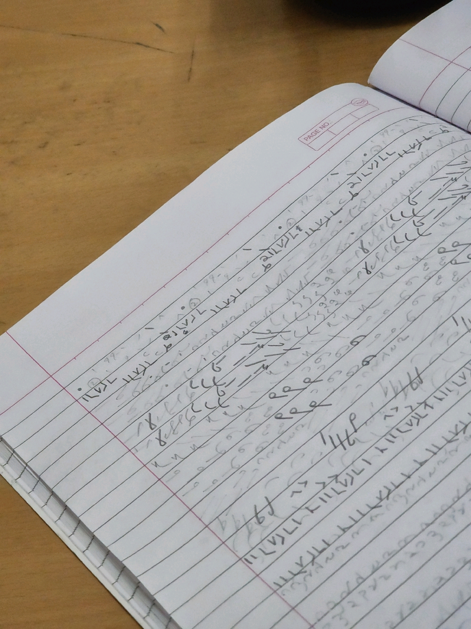 The image shows a close-up, high-angle shot of an open notebook with ruled lines, placed on a wooden surface. The notebook is filled with handwritten shorthand notes, primarily appearing in dark ink. Some lines of shorthand are more legible than others, with symbols, strokes, and dots forming a dense pattern across the pages. A small section of the notebook's cover, featuring a printed "PAGE NO." label and a blank space for numbering, is visible in the upper right portion of the image. The wooden surface beneath the notebook appears to be a desk or table, with a light brown, grained texture. There are no people or specific landmarks of Ajmer, India, visible in the image. The focus is entirely on the notebook and its contents.