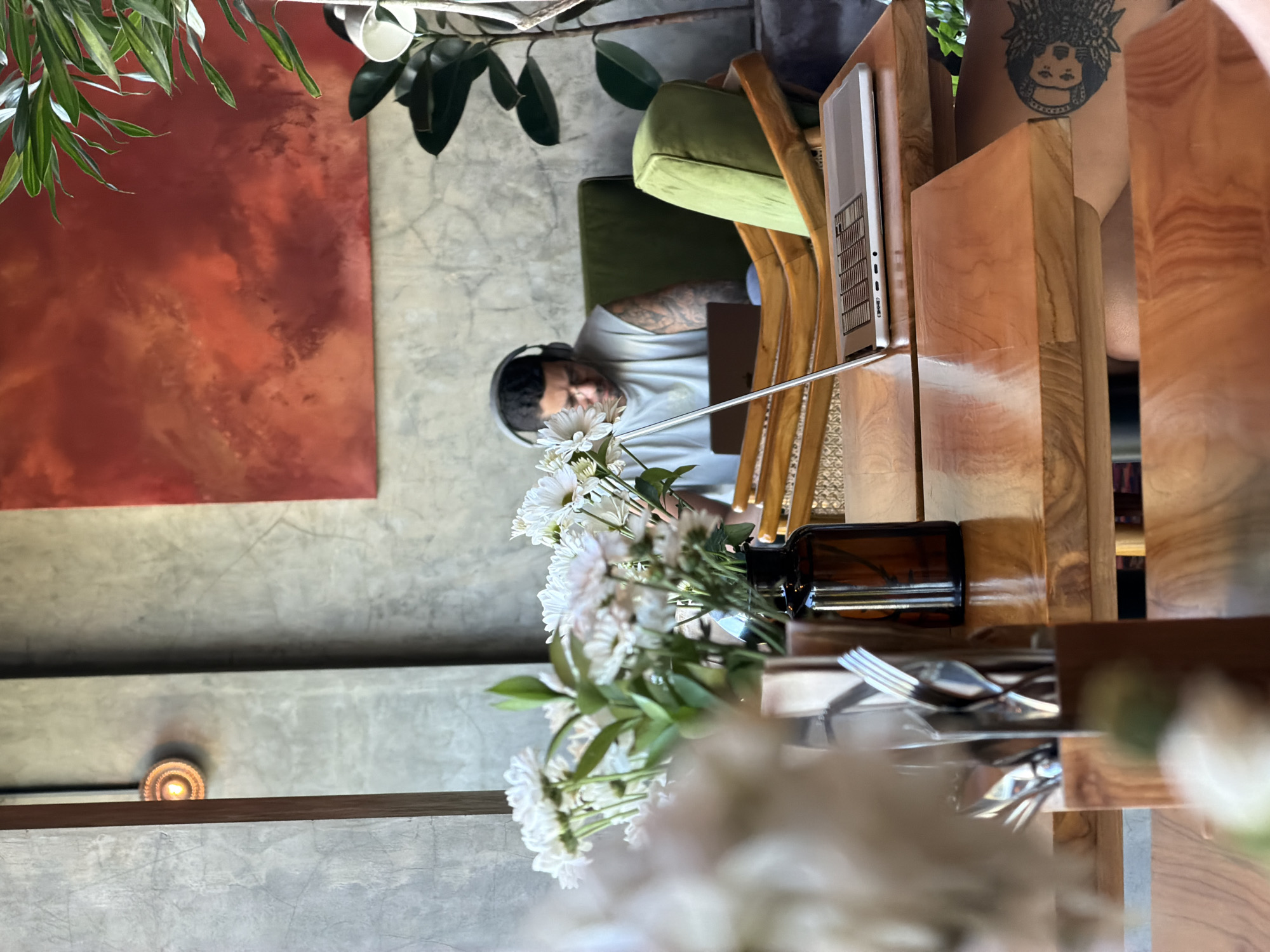 A man wearing headphones is working on a laptop in a cafe in Mengwi, Indonesia. He is seated in a green armchair, and a bouquet of white flowers is in the foreground. The cafe has a modern, industrial aesthetic with concrete walls and wooden furniture. A large abstract painting in shades of red hangs on the wall.