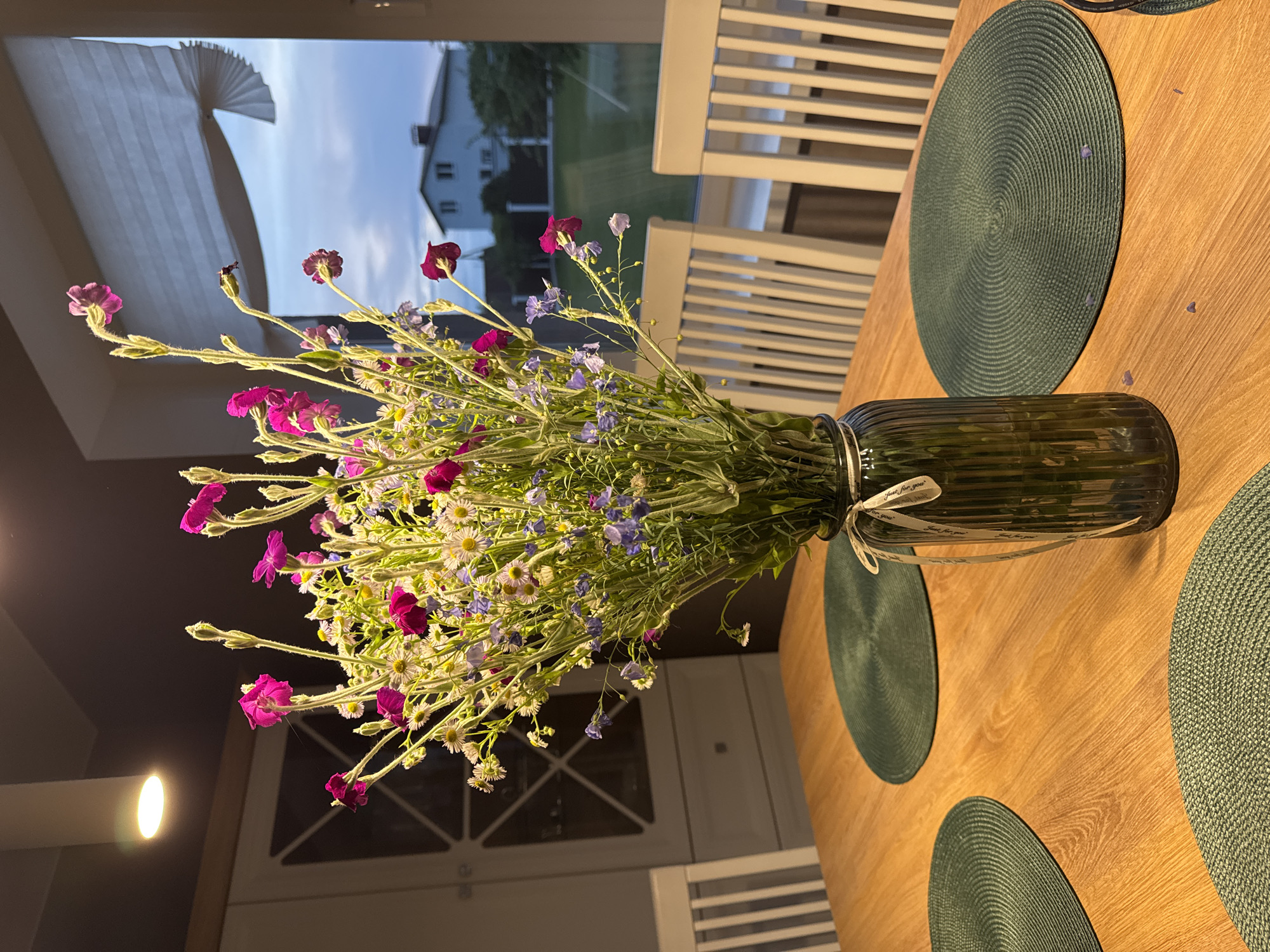 A bouquet of wildflowers, primarily featuring pink tobacco flowers, white daisies, and small blue flowers, is displayed in a tall, ribbed green glass vase on a wooden dining table. The vase is tied with a simple cream ribbon. The table is set with three dark green, woven circular placemats. In the background, through a window, a suburban house and a twilight sky are visible. White wooden chairs are arranged around the table. The scene is illuminated by an overhead light fixture and the natural light from the window. The overall setting appears to be a residential interior, likely a dining room, in Бараўлянскі сельскі Савет, Belarus.