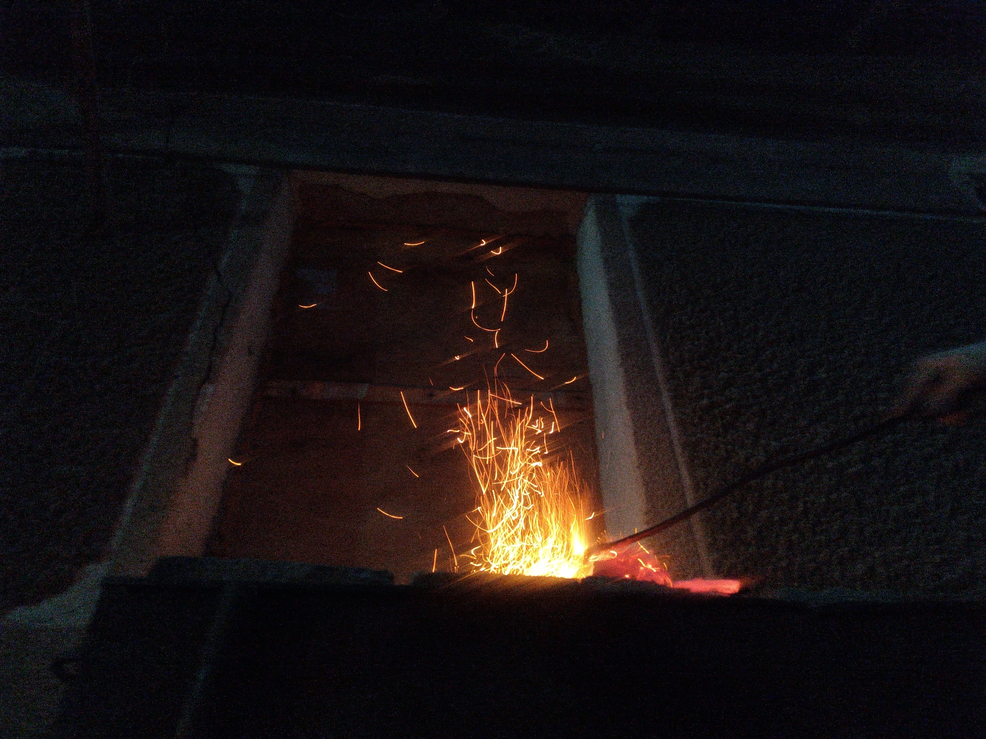 The image depicts a blacksmith's forge at night. A piece of metal, likely being worked on, glows a bright red-orange in the foreground. Above it, a vigorous shower of sparks erupts, illuminating the immediate area with a warm, intense light. The sparks are streaks of yellow and orange against the dark background, suggesting a hot, energetic process. A dark, angled metal rod, held by an unseen hand (partially visible on the right edge), is engaged with the glowing metal, indicating an active forging operation. The background consists of dark, textured surfaces, possibly stone or rough concrete, with a recessed area where the forge appears to be situated. The overall scene is one of intense heat, light, and manual labor, captured in the darkness of night. This activity is occurring at an unidentified blacksmith workshop within the city of Shymkent, Kazakhstan.