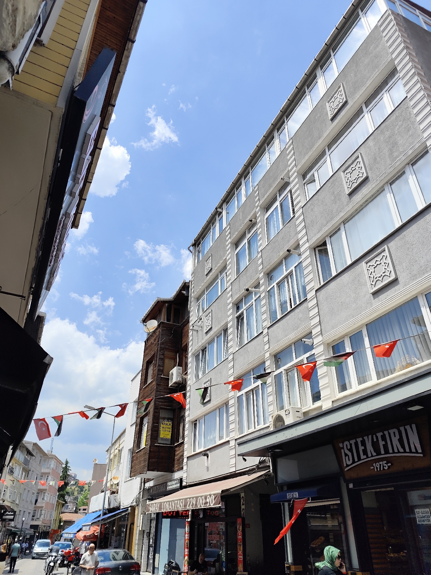 The photograph depicts a street scene in Sarıyer, Turkey, under a partially cloudy sky. The view is looking down the street and a cluster of buildings lines the street. A multistory building dominates the right side of the frame, exhibiting a gray facade with symmetrical, decorative square accents. Windows are numerous and reflect the sky. A banner of flags with red and black markings stretches across the street.
Below the flags on the right, the storefront of "İSTEK FIRIN -1975-" is visible, with a woman wearing a green headscarf standing nearby, appearing to be on her mobile phone.
On the left side, various shops line the street, with signs and awnings. Several people are walking on the sidewalk, and cars are parked along the curb, creating a busy urban environment. The street is narrow, with buildings close together. The image suggests a commercial area within Sarıyer.