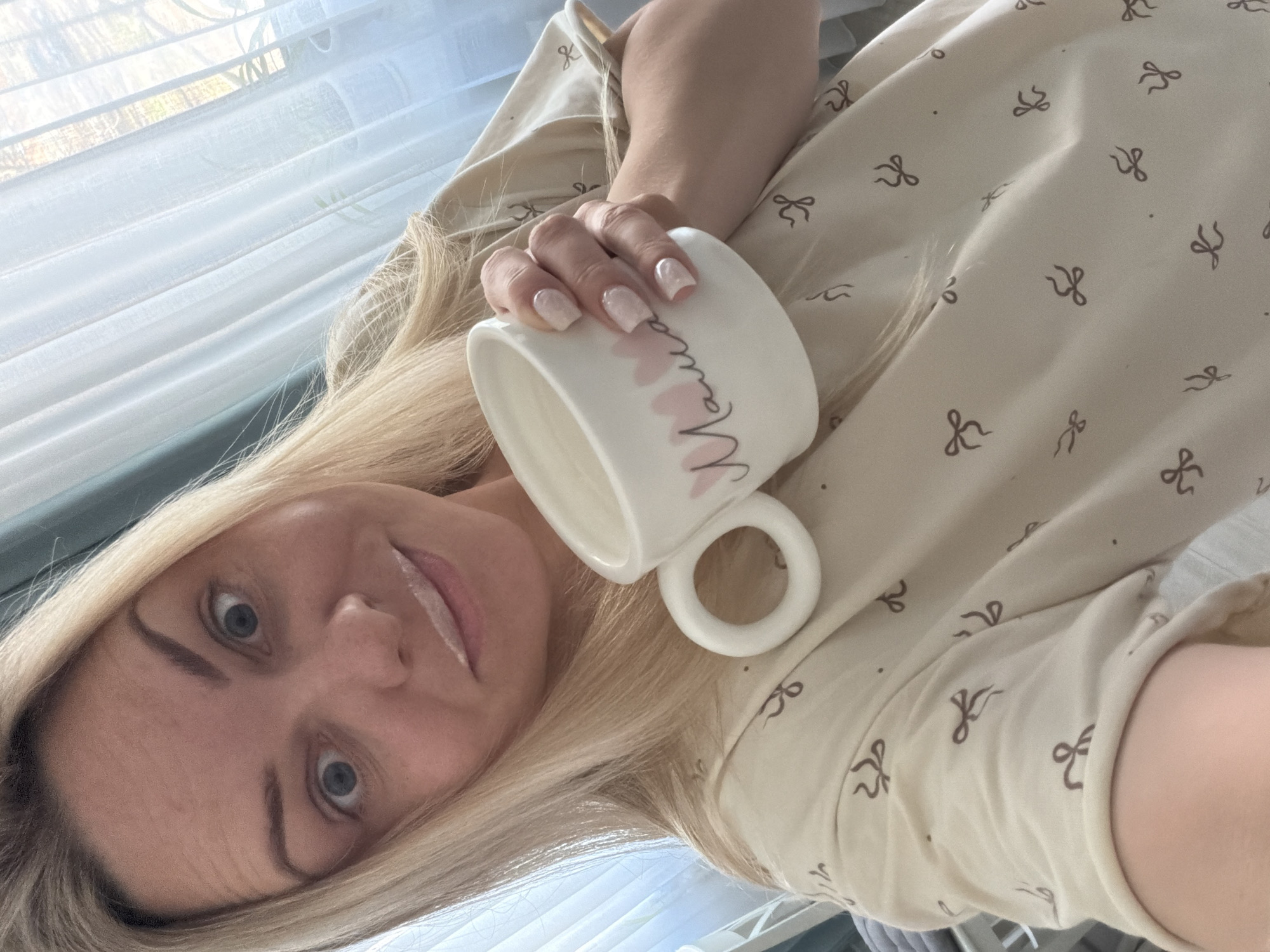 A blonde woman is captured in a rotated, close-up selfie in Votkinsk, Russia, looking directly at the camera with blue eyes and a subtle, soft smile. Her long, straight blonde hair cascades around her face and shoulders. She holds a white ceramic mug with her right hand, showcasing square-shaped nails painted with a light, possibly iridescent, polish. The mug is adorned with pink hearts and the cursive word "Mama" in a reddish-brown or dark pink font. She is wearing a light beige or cream-colored garment, likely pajamas or loungewear, featuring a repeating pattern of small dark brown bow ties and dots. In the background, a window with white horizontal blinds is visible, letting in soft, diffused natural daylight. Through the slightly open blinds, blurry hints of green and brown foliage suggest an outdoor view. The image conveys a serene and relaxed atmosphere, likely taken during the daytime.