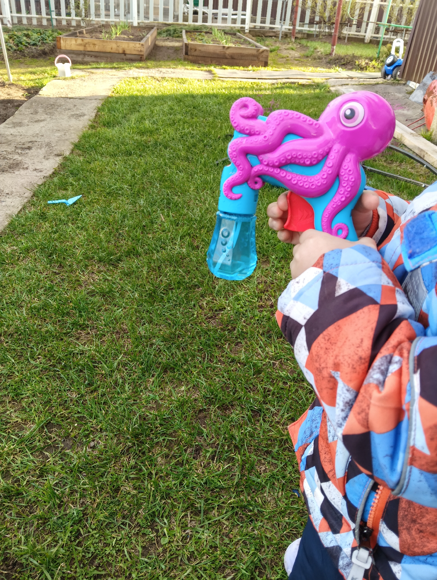 The image presents a close-up, high-angle view of a child's hands holding a colorful toy in a sunny garden in Chaikovsky, Russia. The child, whose upper body and face are not visible, is wearing a vibrant jacket with a geometric pattern in shades of orange, blue, black, and white. They are holding a distinctive pink and blue octopus-shaped bubble gun, which features an attached clear blue container, likely for bubble solution.
The setting is an outdoor garden or backyard. The foreground and midground are covered by green grass, with a grey concrete path running along the left side. A small blue plastic shovel or trowel lies on the grass near the path. In the background, a white wooden picket fence marks the boundary of the garden, behind which two wooden raised garden beds contain small, green plants. A white bucket is visible near the garden beds, and further in the distance, a small blue and white ride-on toy car is parked. Another darker fence structure can be seen in the far background.
The time of day appears to be daytime, with bright sunlight casting shadows on the ground, suggesting clear, sunny weather. The scene conveys a sense of playful outdoor activity in a domestic garden environment. There is no visible text in the image.