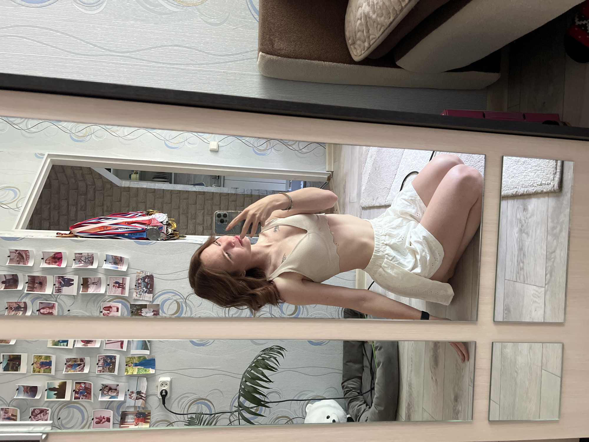 A woman is reflected in a mirror. She has brown hair and is wearing a beige top and white shorts. She is holding a phone and appears to be taking a picture of herself. The mirror reflects a room with patterned wallpaper, framed pictures, a houseplant, and other domestic objects. Based on metadata, the location is in Chaikovsky, Russia.