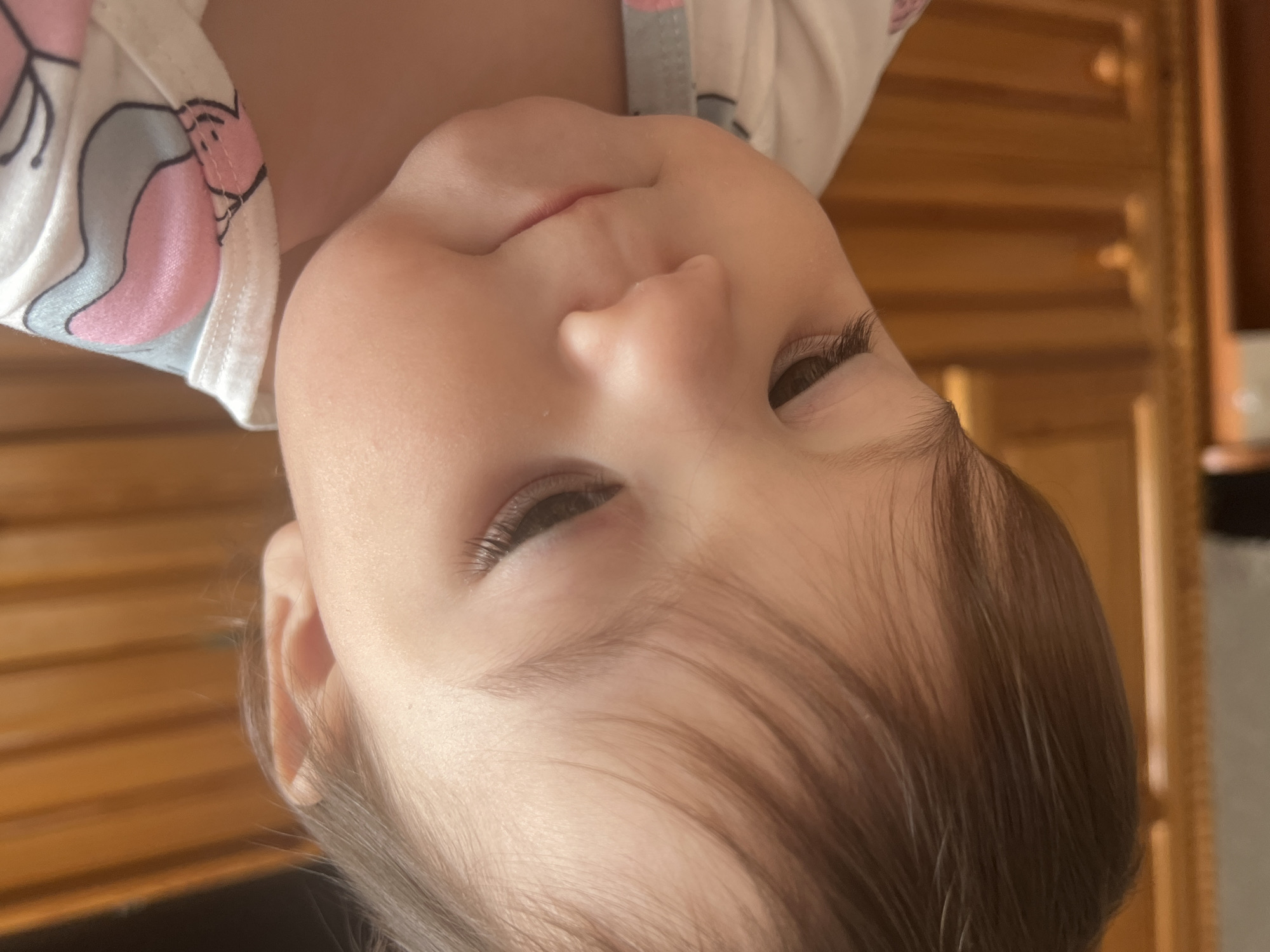 Close-up portrait of a baby, possibly Caucasian, with fair skin, brown hair, and long eyelashes. The baby is wearing a white garment with a pink and gray cartoon design. The background appears to be a wooden wall or cabinet. The image is rotated, making the baby's face appear upside down. The setting does not show any recognizable feature related to the city of Elena, Bulgaria.