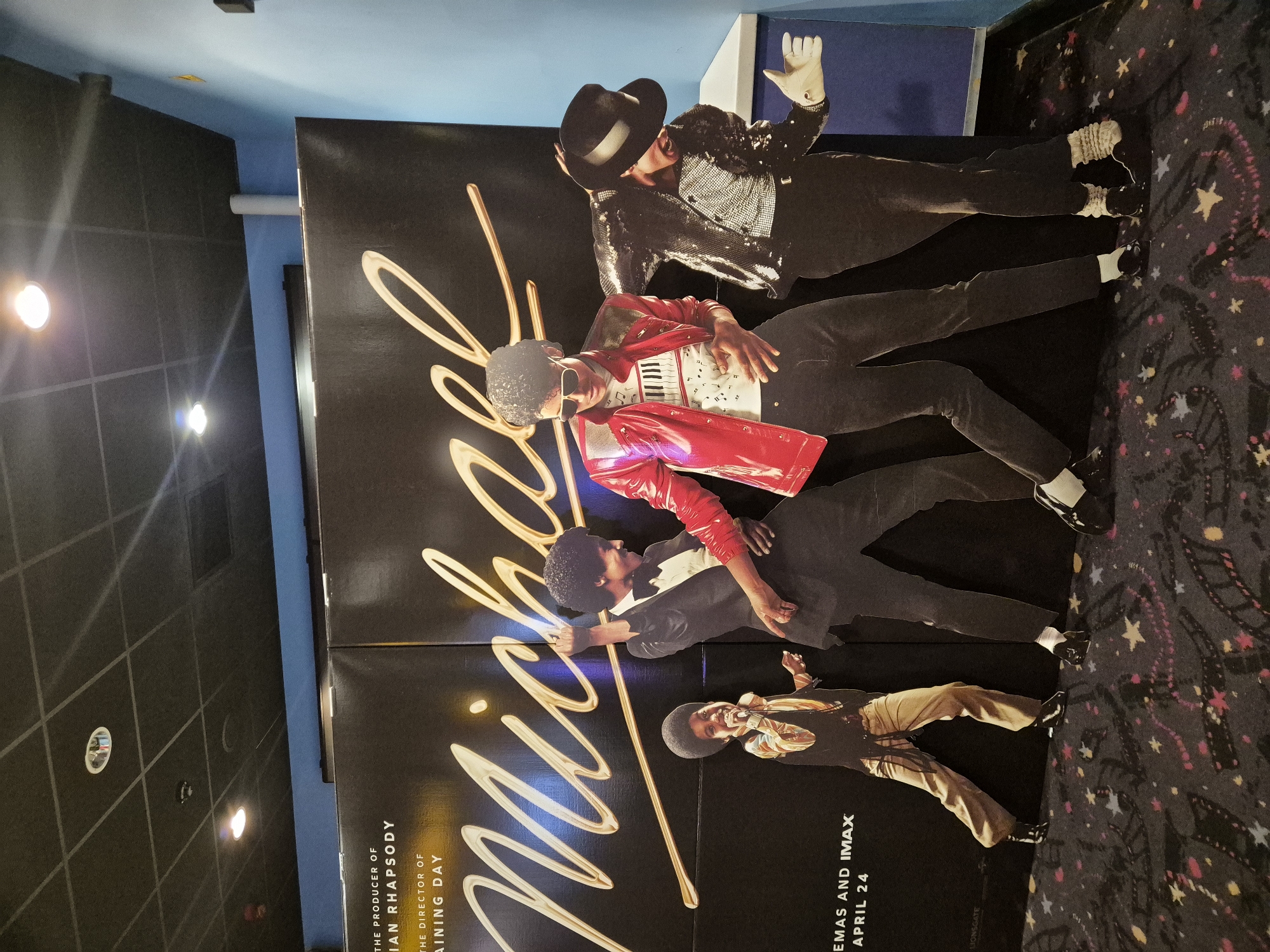 The image shows a movie poster for the film "Michael," apparently displayed in a cinema location. The poster features multiple images of Michael Jackson in different outfits and poses. The text on the poster includes the title "Michael" in large, stylized letters, along with the information "THE PRODUCER OF IAN RHAPSODY," "THE DIRECTOR OF RAINING DAY," and "CINEMAS AND IMAX APRIL 24." The poster is black with gold-colored lettering and accents. The background environment shows a dark, carpeted floor and a blue wall. The location in the city of West Suffolk, United Kingdom is not explicitly stated, but this level of detail is beyond the scope of the scene.
