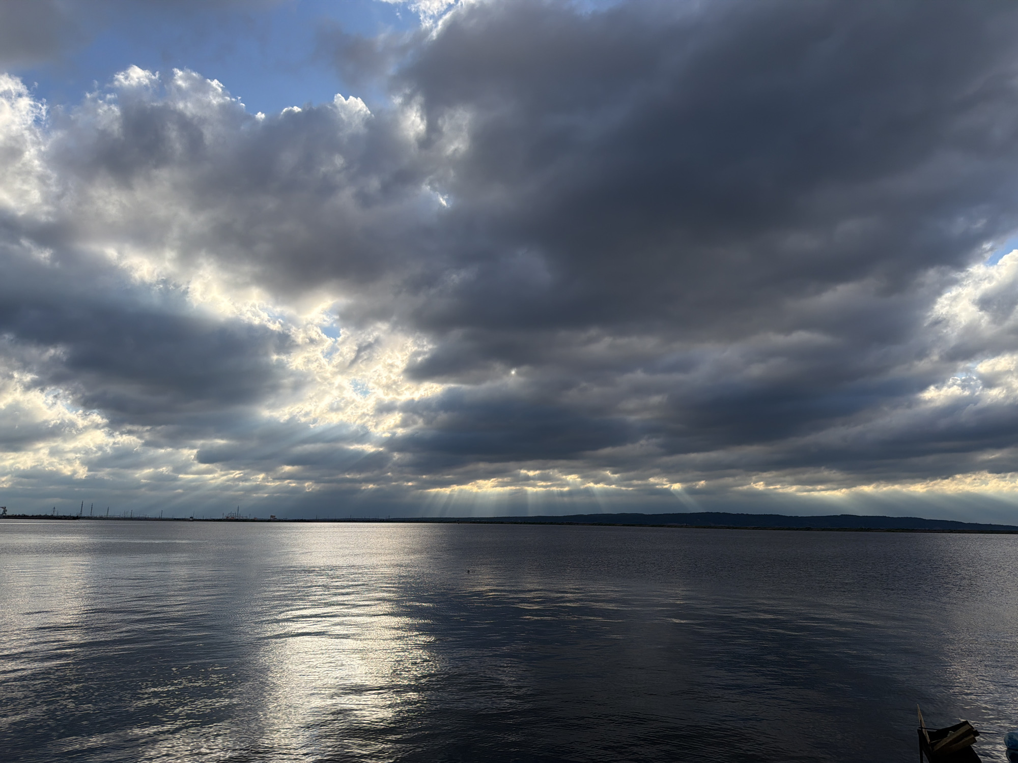 The image depicts a wide body of dark, rippling water under a dramatic sky filled with heavy, dark clouds. Rays of light pierce through gaps in the clouds, illuminating sections of the water with a bright, reflected sheen and casting beams downwards. The horizon is a dark, indistinct line, suggesting land or distant structures, possibly industrial in nature, with vertical elements like poles or towers visible against the sky in the distance. A small wooden boat is partially visible in the lower right corner. The scene evokes a sense of moody, atmospheric weather over a large expanse of water, possibly a reservoir or river, consistent with the location of Chaikovsky, Russia, known for its proximity to the Kama River and associated hydroelectric dam.