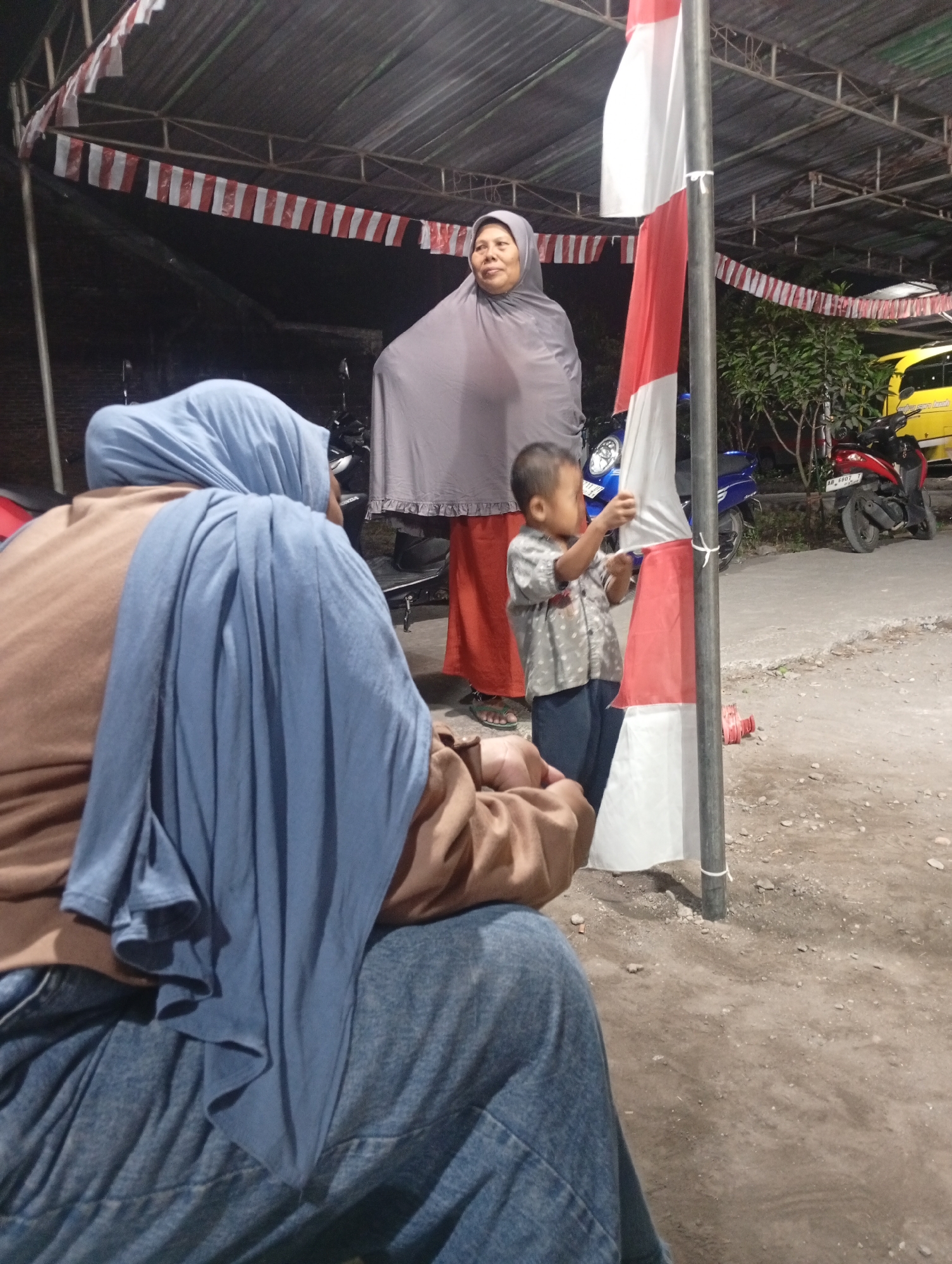 In Sewon, Indonesia, a group of people are gathered under a tented structure at night. In the foreground, a person wearing a blue hijab and brown top sits with their back to the camera, while another individual in a grey hijab stands to the right. A young child in a patterned shirt and dark pants is looking at a red and white flag attached to a pole. In the background, motorcycles and a yellow vehicle are visible, along with a string of red and white flags adorning the tent. The ground is a mixture of dirt and gravel.