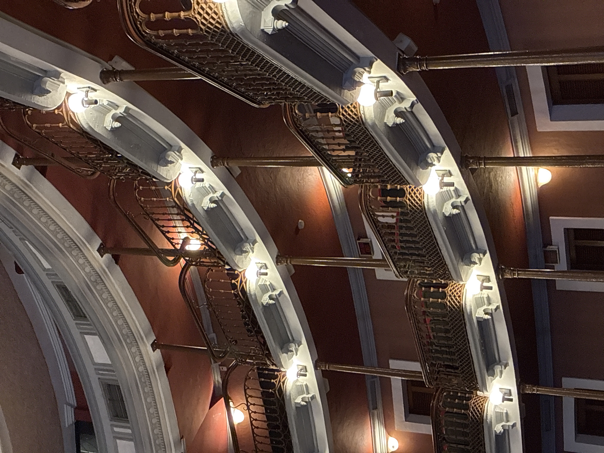 The image presents an interior view of a building, likely taken from a lower level looking upwards. The architectural details are prominent, featuring a series of arches constructed with a combination of white, grey, and possibly metallic-colored elements. These arches are adorned with ornate details, including decorative moldings and what appear to be lighting fixtures spaced along their length. Hanging beneath each arch is a grid-like structure, which may be a form of ventilation or decorative screening.
The background wall is painted in a dark reddish-brown hue, accented with white or light-colored trim. Light sources are visible as glowing orbs, presumably illuminating the scene. The location of the building within the city of Mazatlán, Mexico is not evident from the image.