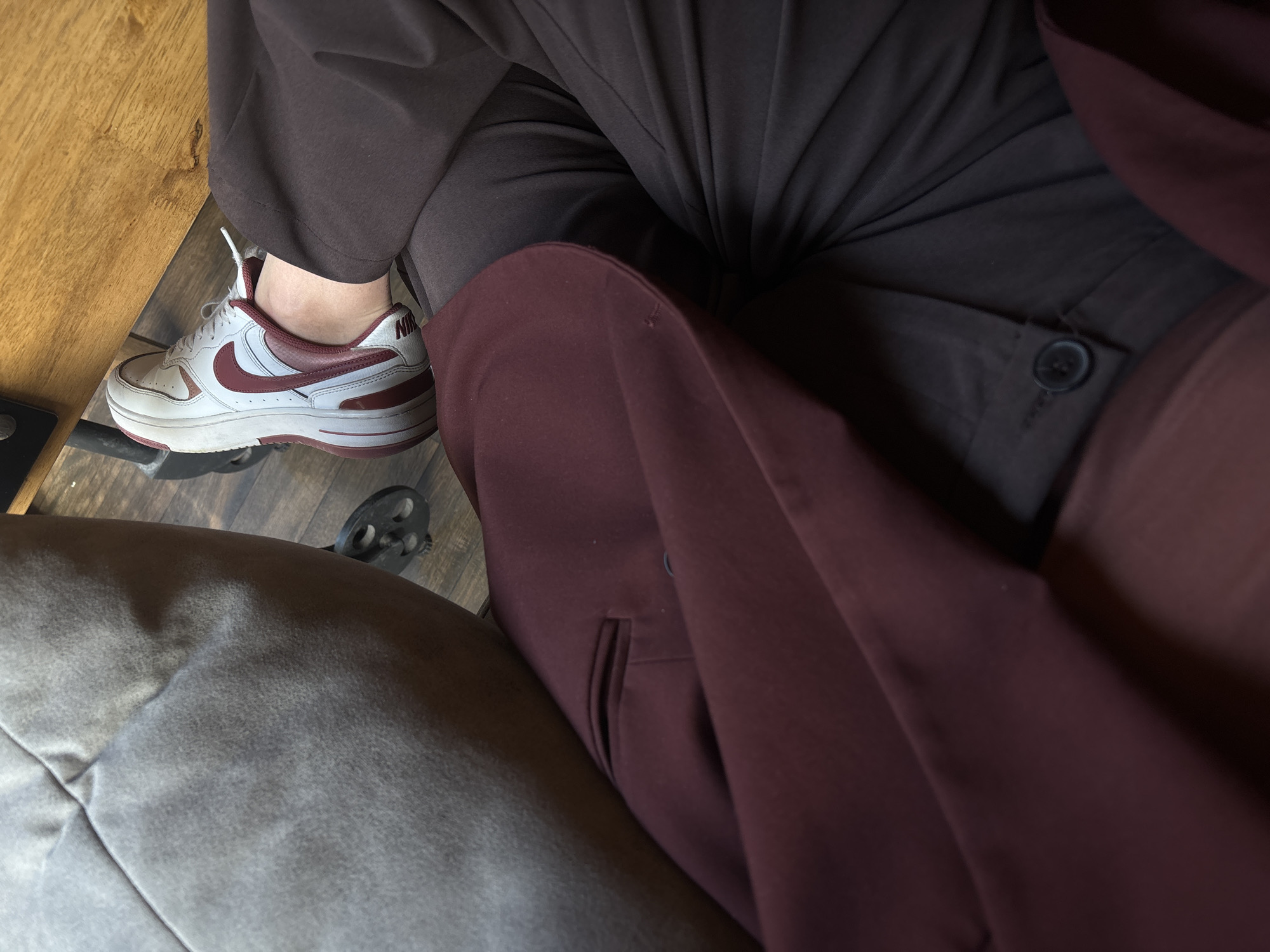 This image shows a seated person wearing gray sweatpants, brown pants, and a maroon coat. The person is wearing a white Nike shoe with a burgundy swoosh. The environment includes a wooden surface on the left and the legs of a chair on the floor. The location cannot be determined from the image.
