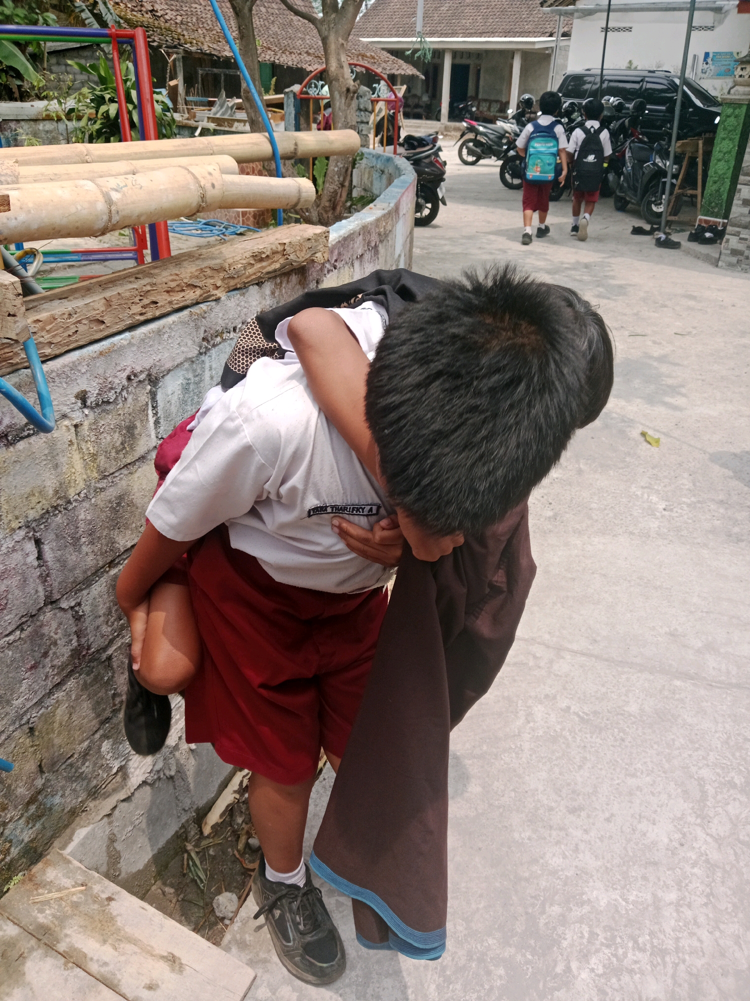 This image depicts a street scene in Salatiga, Indonesia, likely near a school given the presence of students in uniform. In the foreground, a young boy wearing a white polo shirt and red shorts is carrying another younger child on his back. The younger child is wearing a dark fabric draped over their body and is partially obscuring the older boy's face and torso. The older boy's left leg is bent, and his foot is visible, wearing a black shoe and white sock. He is leaning against a textured, light-colored wall.
In the background, two other young boys in similar school uniforms are walking away from the camera, each carrying a backpack. The boy on the left has a light blue backpack, and the boy on the right has a black backpack. They are walking on a concrete pathway. Several motorcycles are parked to the right of the pathway, and a black vehicle, possibly an SUV or minivan, is parked behind them. The environment appears to be outdoors with a building in the background, and some vegetation and playground equipment are visible on the left side of the image. The overall lighting suggests it is daytime, with shadows cast on the ground.
