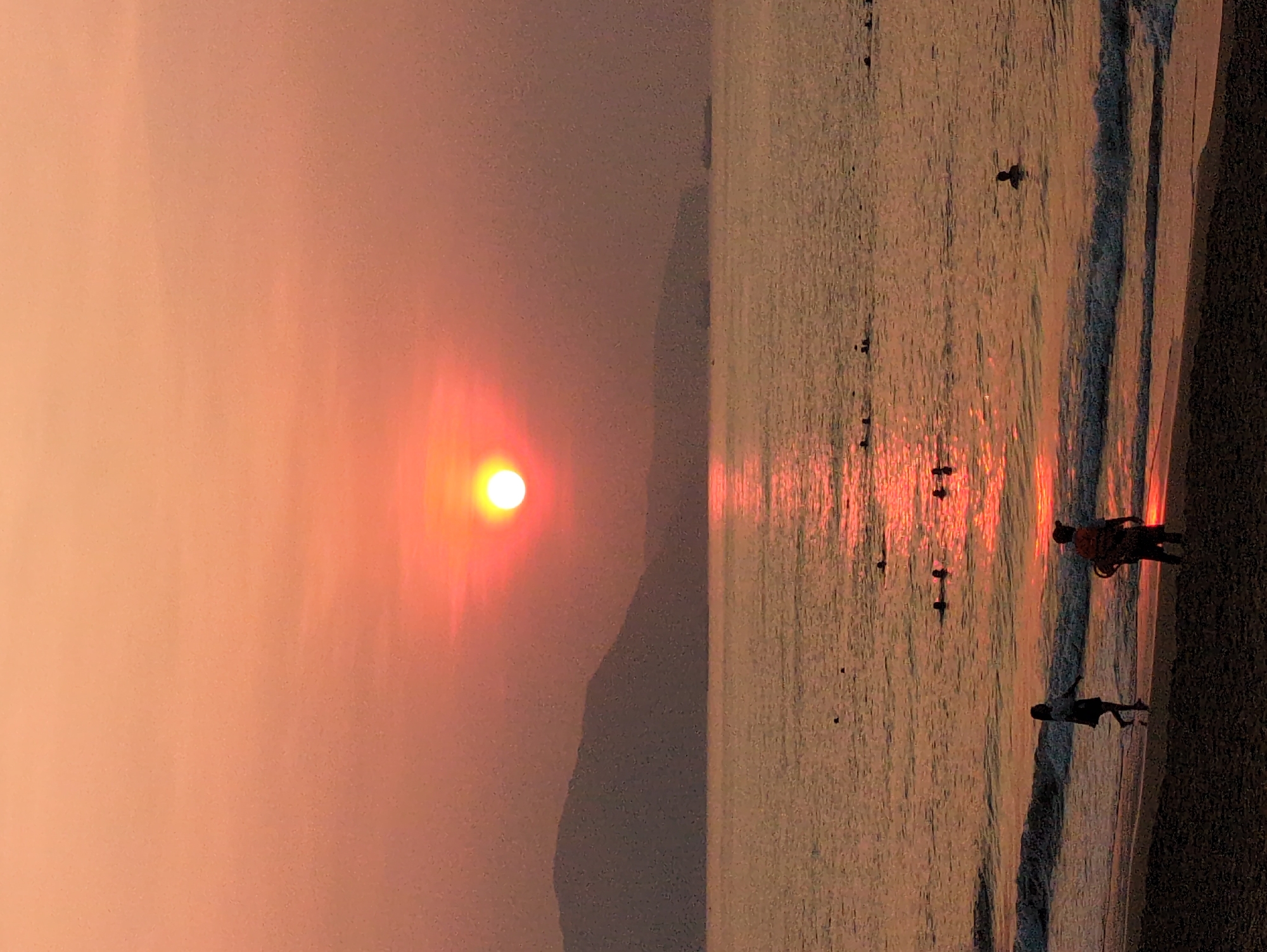 The image depicts a beach scene at South Quy Nhon Ward, Vietnam, during a sunset. The sky is a gradient of orange and red, with the sun visible as a bright orb over a land mass in the distance. The sea reflects the colors of the sunset, with a shimmering path of light extending from the sun towards the shore. Several individuals are in the water and on the beach, their silhouettes visible against the light. The shoreline is defined by gentle waves, and the beach itself is dark. The photograph is oriented vertically.