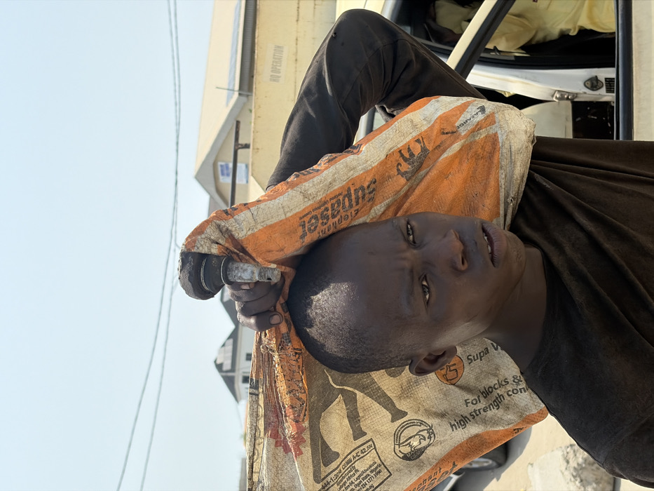 An outdoor, eye-level portrait shows a young male subject in Jakande, Nigeria, illuminated by strong daylight under a clear sky. The subject, with dark skin and short dark hair, wears a dark-colored shirt. He carries a large, torn, woven polypropylene sack, identifiable as an "Elephant Supaset" brand cement bag with orange and beige coloring and text "For blocks & high strength concrete," across his left shoulder and head. His right hand grips a dirty, metallic automotive component, possibly a ball joint or suspension part, which protrudes from the bag and rests against his head. His expression is unsmiling, with his gaze directed slightly to his right. In the background, under overhead utility wires, stand residential buildings with light-colored roofs, some equipped with solar panels. Further back, a larger building displays "HQ OPERATION" on its side. A white vehicle with an open passenger door is partially visible on the far right.
