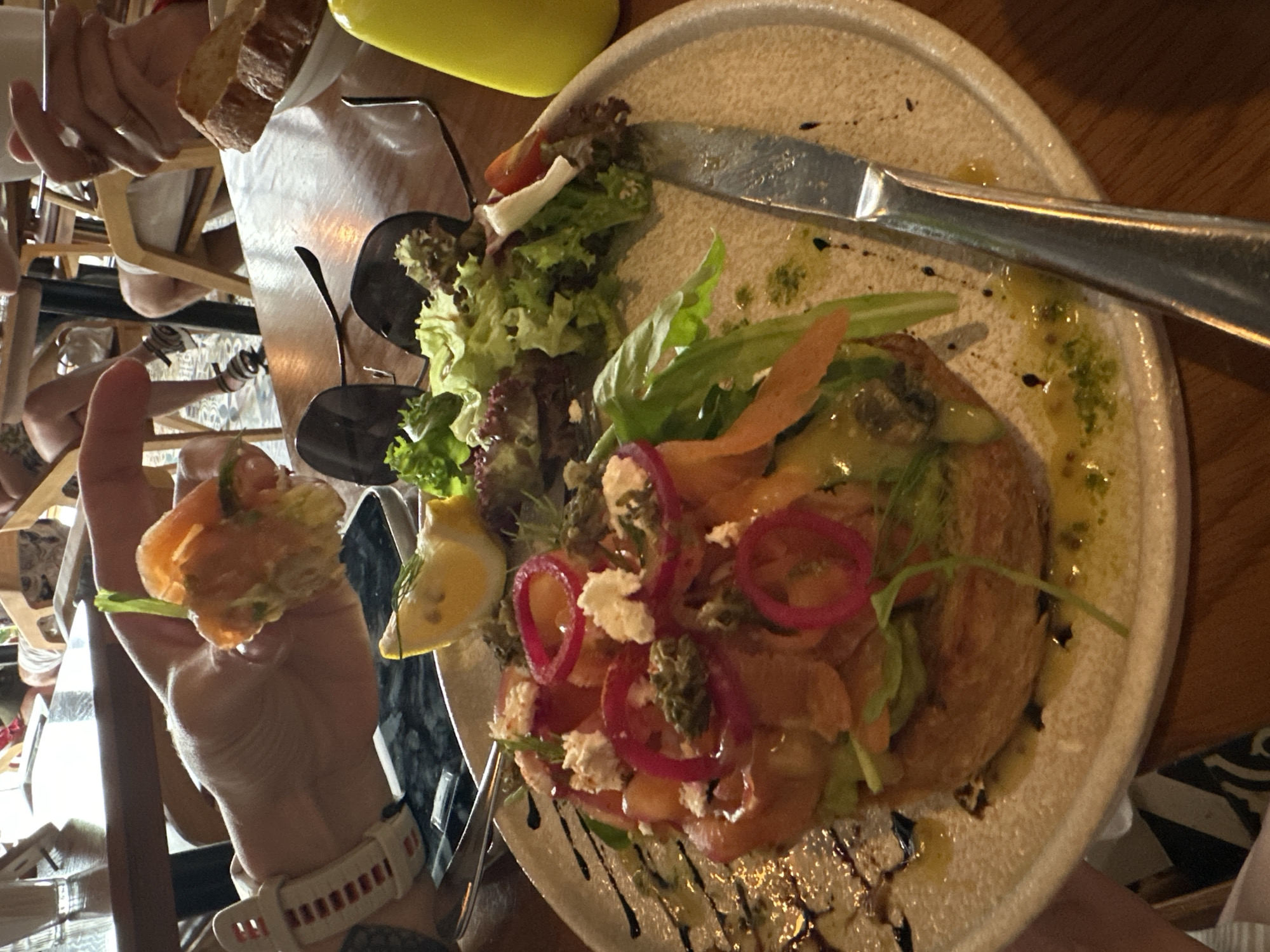 The image shows a meal at an unidentified restaurant in Nha Trang Ward, Vietnam. A plate dominates the right half of the image, containing a dish topped with salmon, red onions, a white cheese-like substance, and an assortment of greens. A lemon wedge is visible on the left side of the plate. Balsamic glaze is drizzled on the plate. A stainless-steel knife rests on the plate. A hand on the left of the image is holding a small portion of the meal. A forearm is visible wearing a white watch with red detailing. In the background are a set of glasses, part of a yellow bottle and what appears to be other diners.
