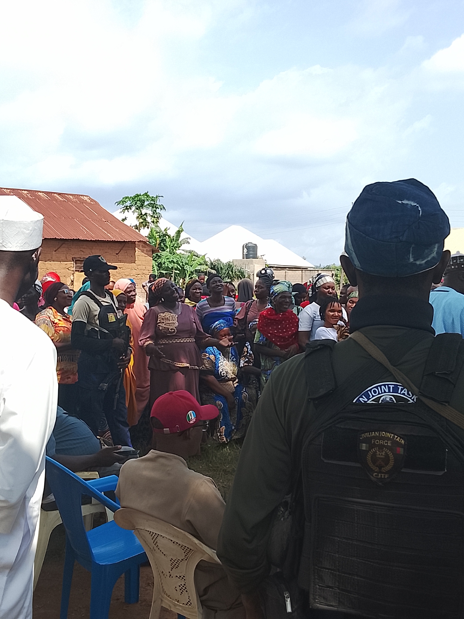 The image depicts an outdoor gathering of people in what appears to be a village setting, with humble dwellings and a cloudy sky. The scene is populated by a diverse group, predominantly women, dressed in colorful traditional attire. Several individuals are looking towards the foreground, while others are engaged in conversation or observing something to their right.
In the foreground, a person wearing a dark blue cap and a tactical vest is the most prominent figure. The vest has patches with visible text, including "CIVILIAN JOINT TASK FORCE" and "CJTF". This suggests a presence of security or a local volunteer force. To the left, a person in a white garment and a white cap is partially visible, facing away from the camera.
The overall atmosphere seems to be one of community interaction, perhaps a local event or a political gathering, given the presence of the uniformed individual and the general assembly of people. The lighting indicates it's daytime, with overcast conditions. The presence of basic structures, possibly homes, and the style of clothing point towards a location in Nigeria, specifically Akwanga. The expressions on the faces of the people in the crowd are varied, ranging from attentive to engaged, and some appear to be smiling. There is a sense of lively communal engagement.