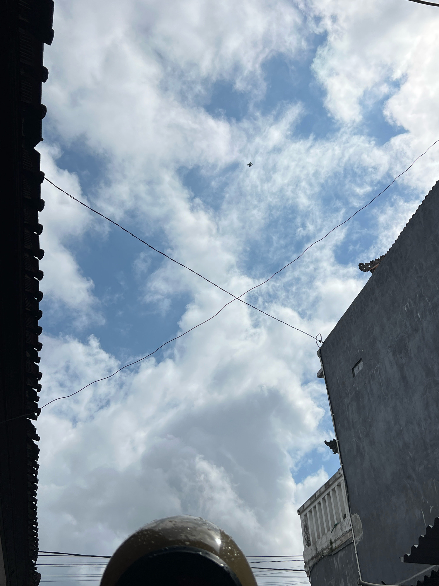 The image, taken from a low angle looking upwards, depicts a sky filled with white, puffy clouds against a bright blue backdrop. Two dark, thin wires are stretched across the frame, intersecting in the center. On the left, the dark, textured edge of a building runs vertically along the frame. On the right, a grey, modern building with a small, dark window and a white balcony railing is visible. A small, dark object, possibly a drone or a kite, is visible high in the sky. At the bottom of the frame, the curved, reflective surface of a helmet is partially visible, suggesting the viewpoint of someone on a motorcycle or scooter. The overall impression is an urban street scene in Denpasar, Indonesia, on a day with variable cloud cover.